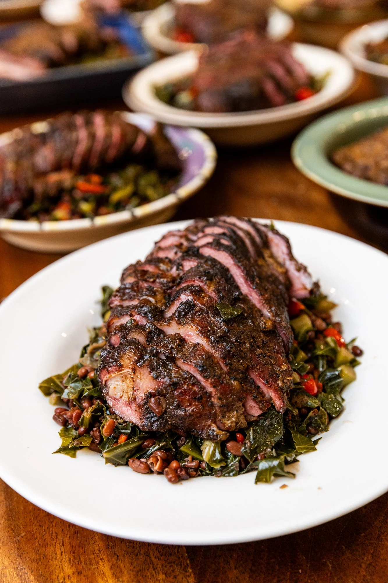 A plate of sliced smoked ham served on a bed of cooked greens and beans, with several dishes of similar food in the background. Southbound Food Festival in Birmingham, Alabama.