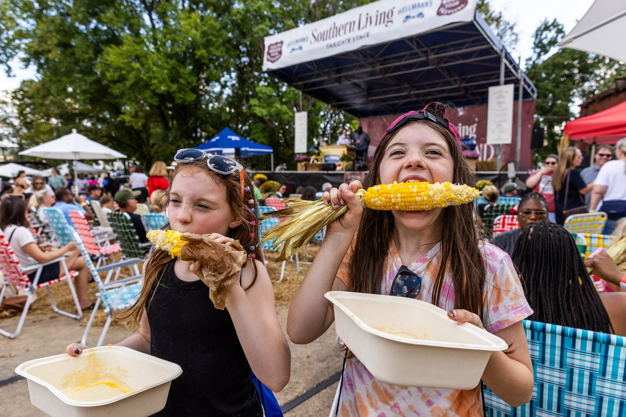 Two young girls with corn cobs eating at an outdoor event with a stage, crowd, and tents in the background. Southbound Food Festival in Birmingham, Alabama.