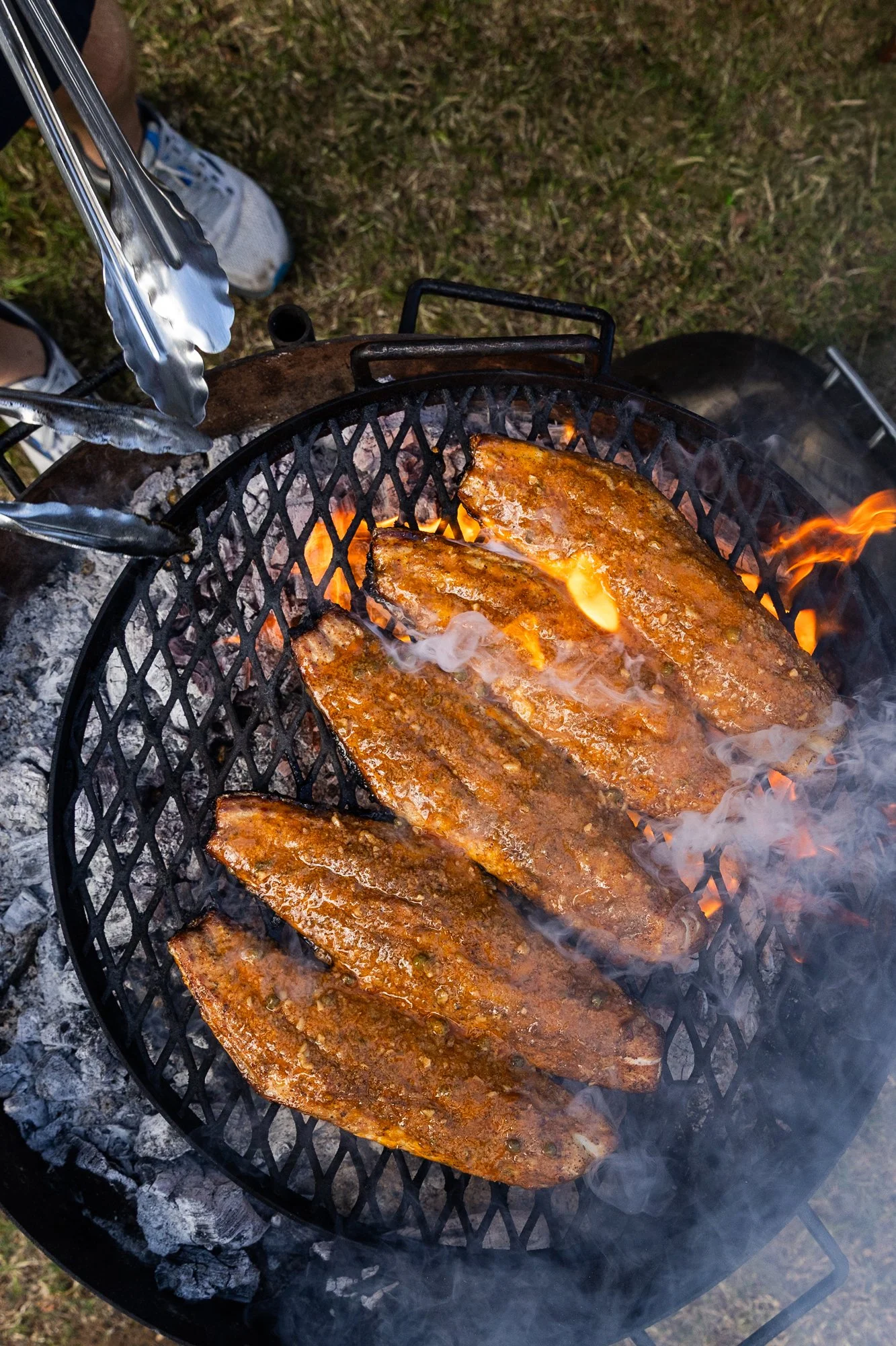Fish fillets grilling on a barbecue over open flames, with tongs visible in the top left corner. Southbound Food Festival in Birmingham, Alabama.