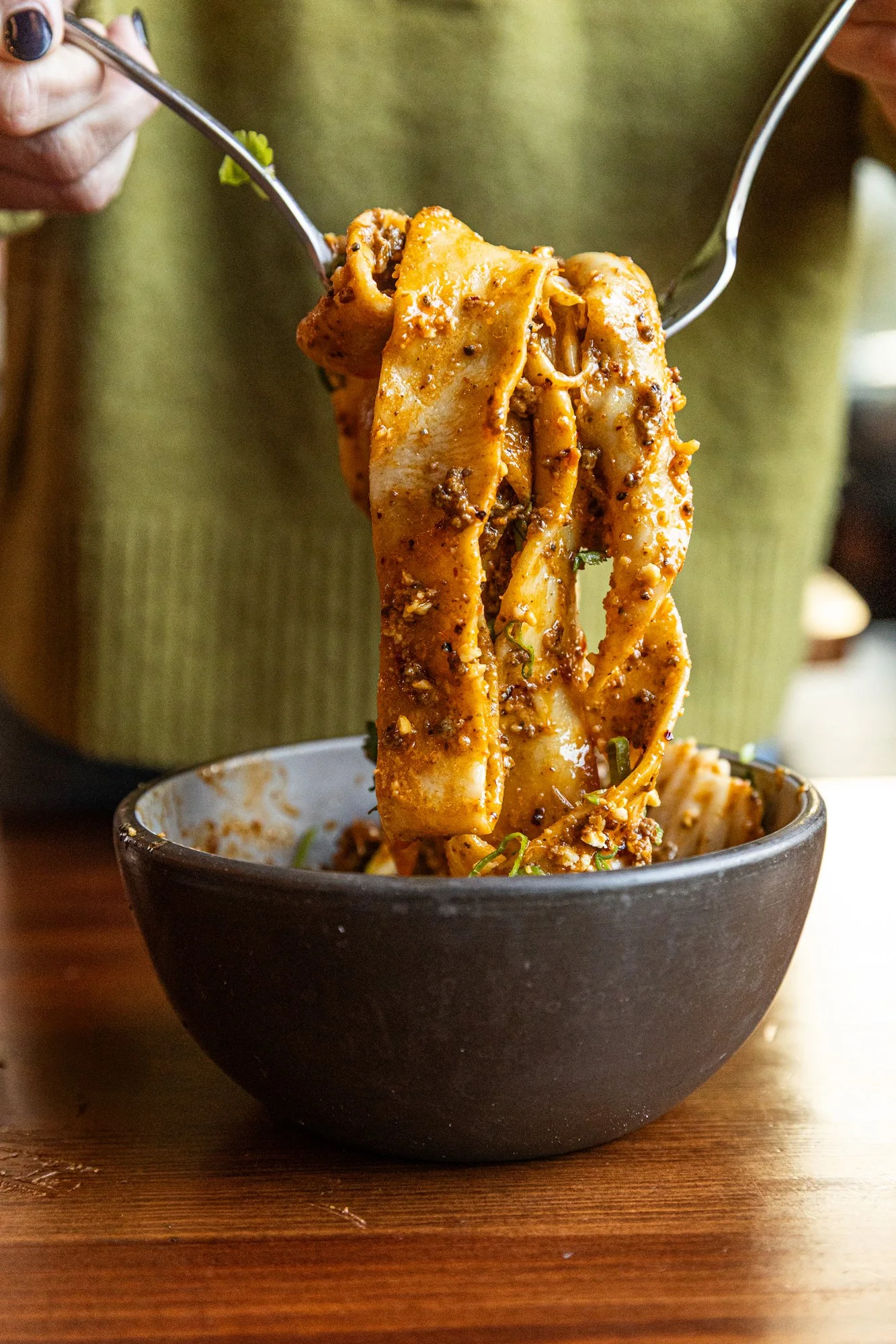 Person holding chopsticks with a bowl of wide flat noodles covered in sauce and spices.