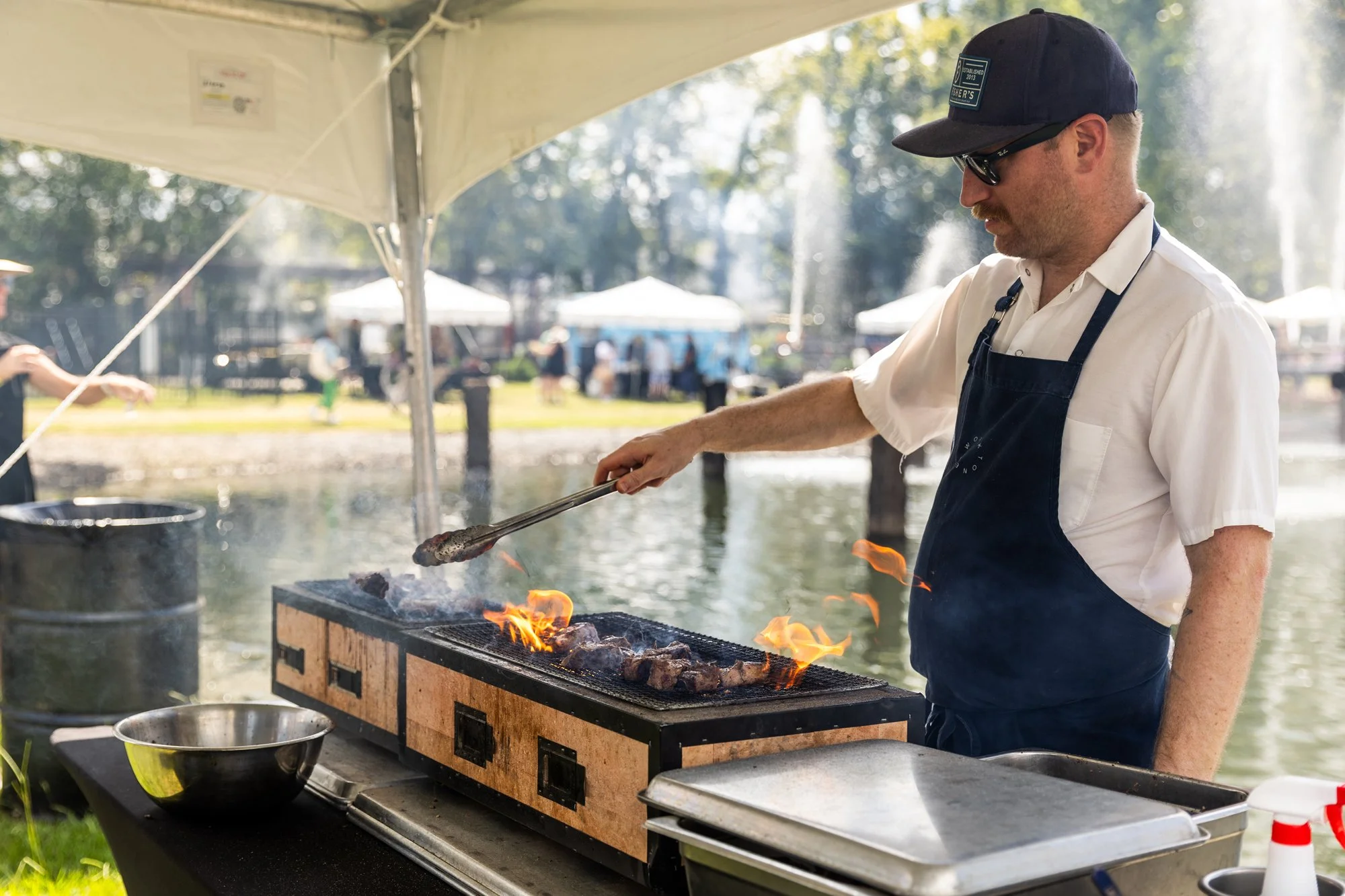 Man in white shirt, black apron, and baseball cap grilling meat on an outdoor barbecue near water with tents and people in the background. Southbound Food Festival in Birmingham, Alabama.