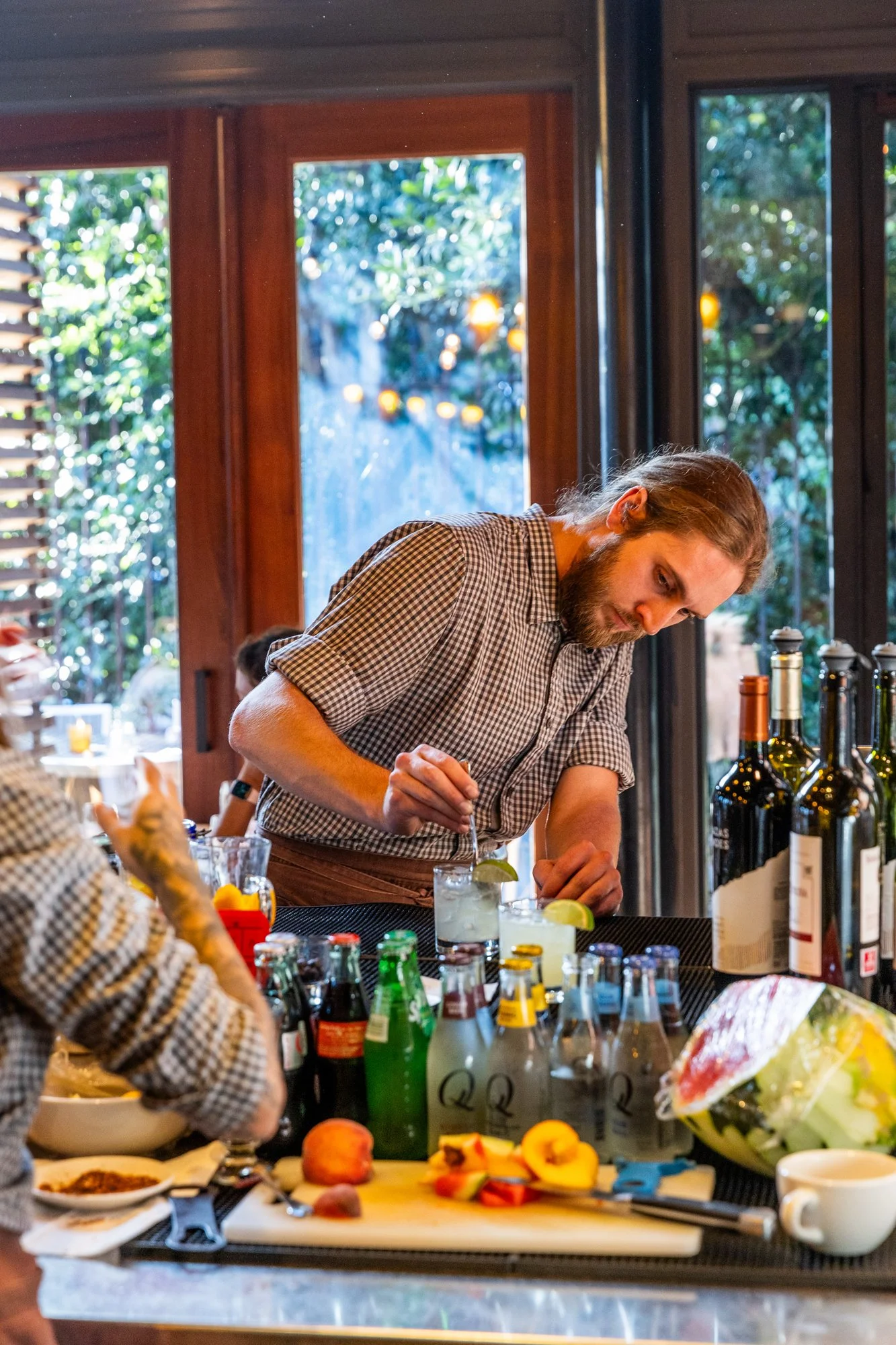 A man preparing drinks at a bar in a restaurant with large windows and greenery outside. Ovenbird Restaurant in Birmingham, Alabama.