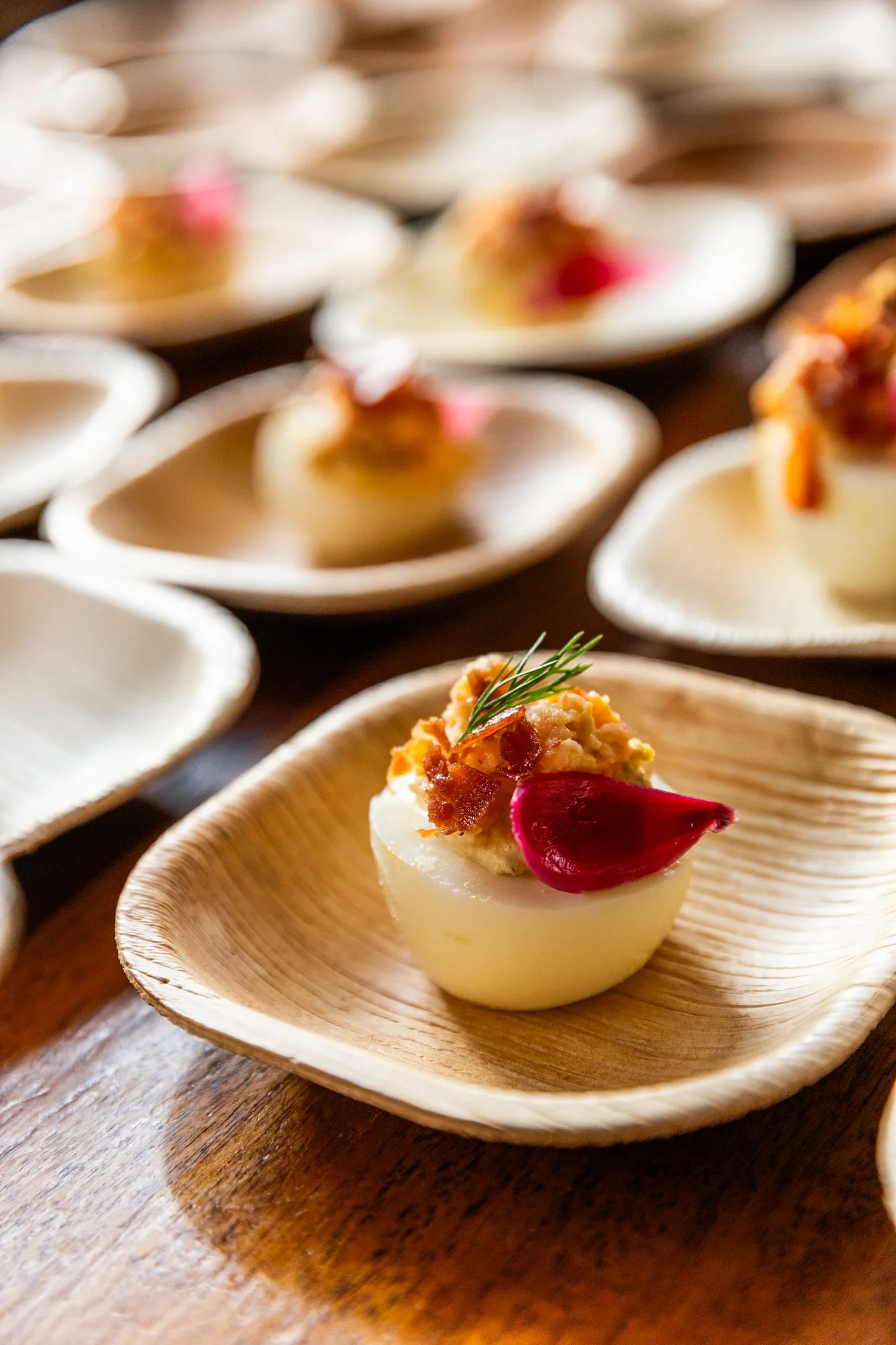 Deviled egg garnished with herb, bacon, and a pickled onion on a wooden dish, with more deviled eggs blurred in the background. Southbound Food Festival in Birmingham, Alabama.