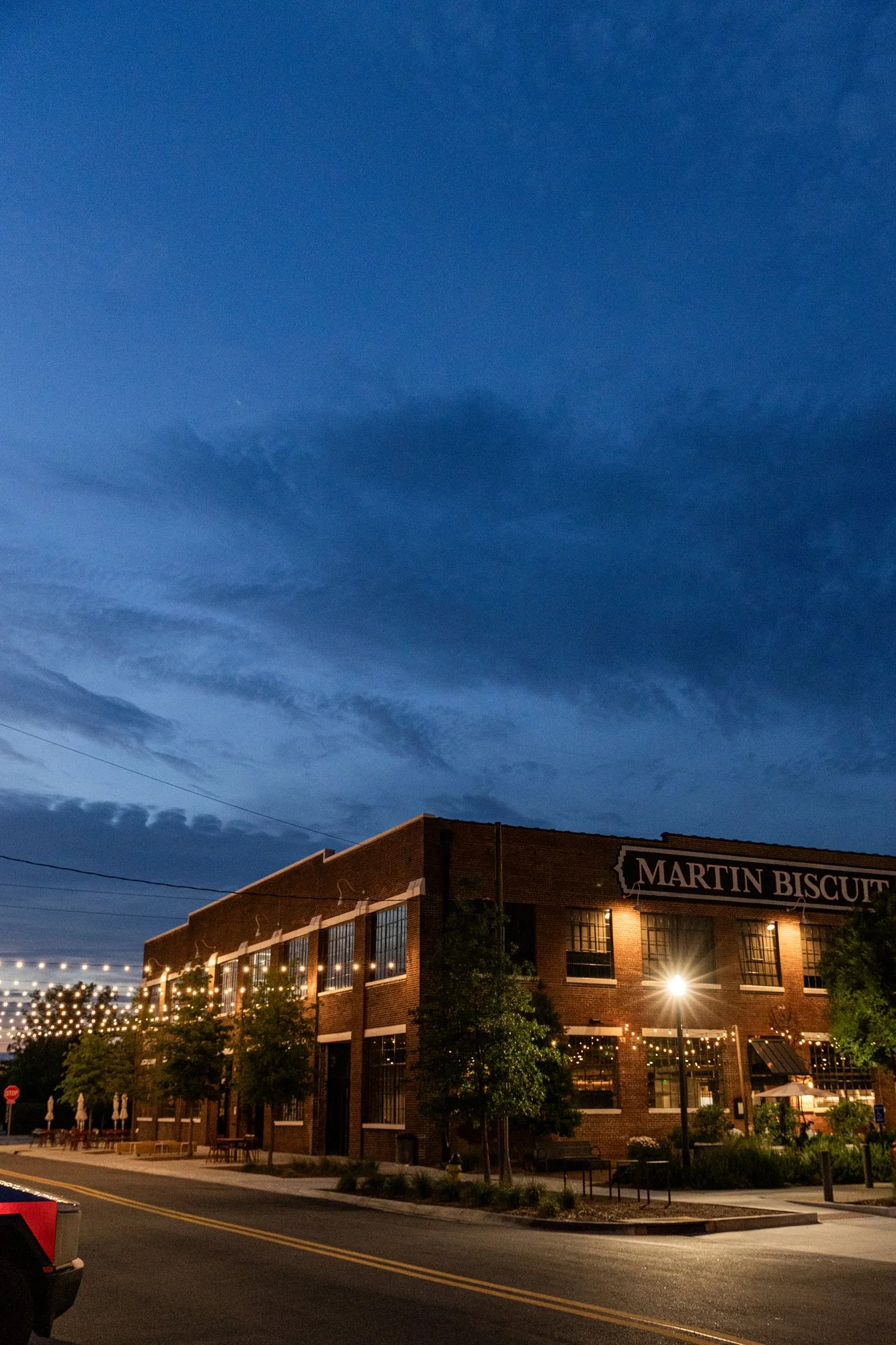Nighttime scene of a brick building with a sign that reads "MARTIN BISCUIT."  Pepperplace, large windows with warm lighting, trees outside, and string lights overhead. Hot andHot Fish Club Restaurant in Birmingham, Alabama.