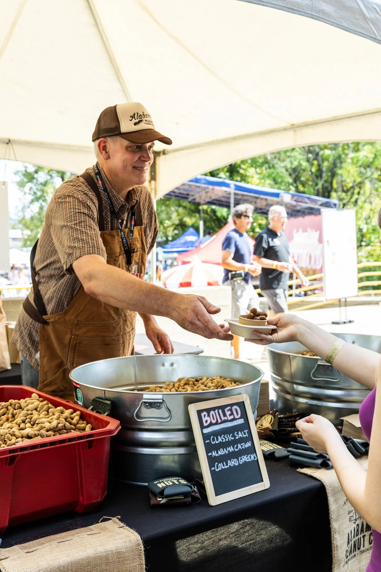 A man in a tan apron and baseball cap is handing a small paper bowl of boiled peanuts to a woman at an outdoor event under a tent. Southbound Food Festival in Birmingham, Alabama.