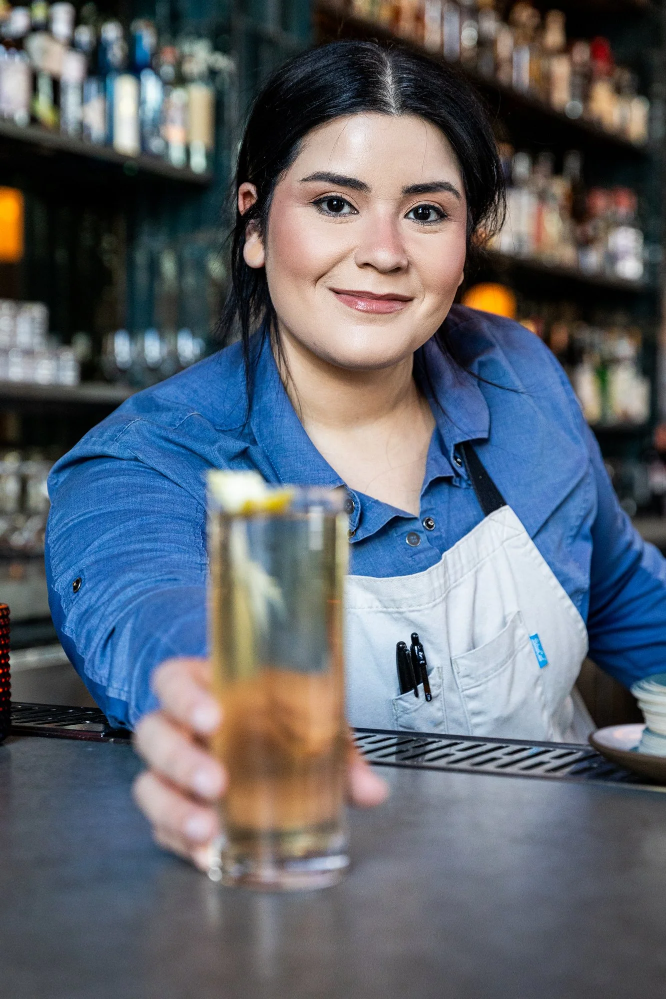 Bartender in a blue shirt and white apron handing a drink to the camera behind a bar.