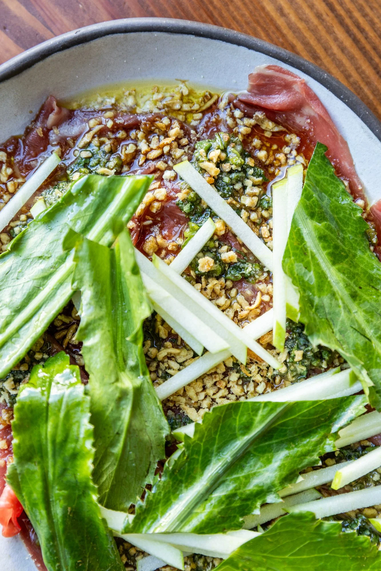Close-up of a dish with sliced raw beef, chopped nuts, green leafy vegetables, and shredded white vegetables in a gray bowl on a wooden surface.