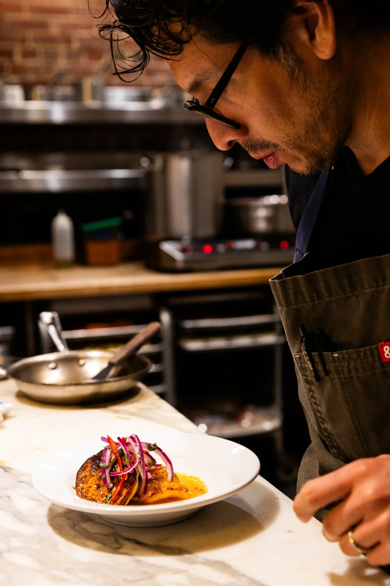 A chef inspecting a plated gourmet dish with meat, topped with colorful shredded vegetables, on a white plate in a professional kitchen. Ovenbird Restaurant in Birmingham, Alabama.
