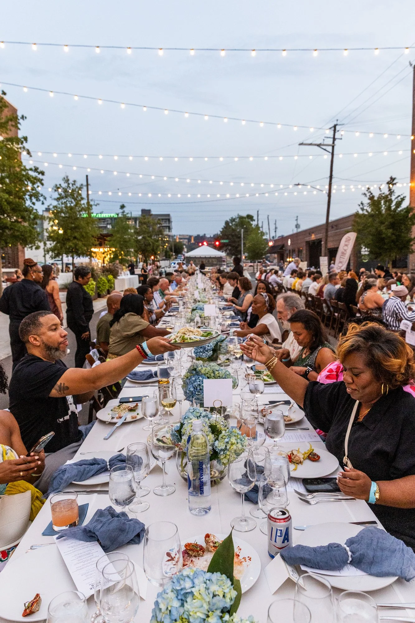 Outdoor dinner party with long table, people serving and dining, string lights overhead, urban street setting with trees and buildings. Southbound Food Festival in Birmingham, Alabama.