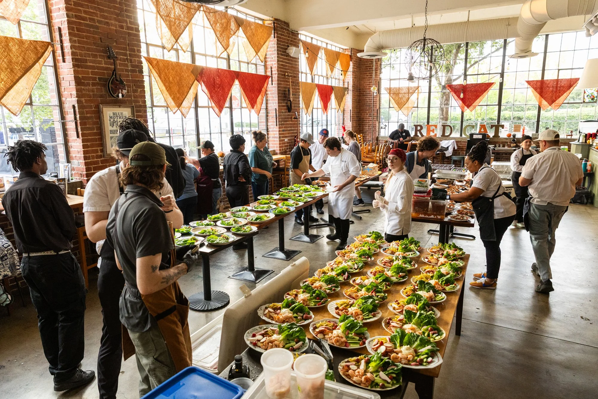 People in a restaurant preparing and serving plates of salads and appetizers for a buffet. Decorated with orange and red bunting flags and large industrial windows. Southbound Food Festival in Birmingham, Alabama.