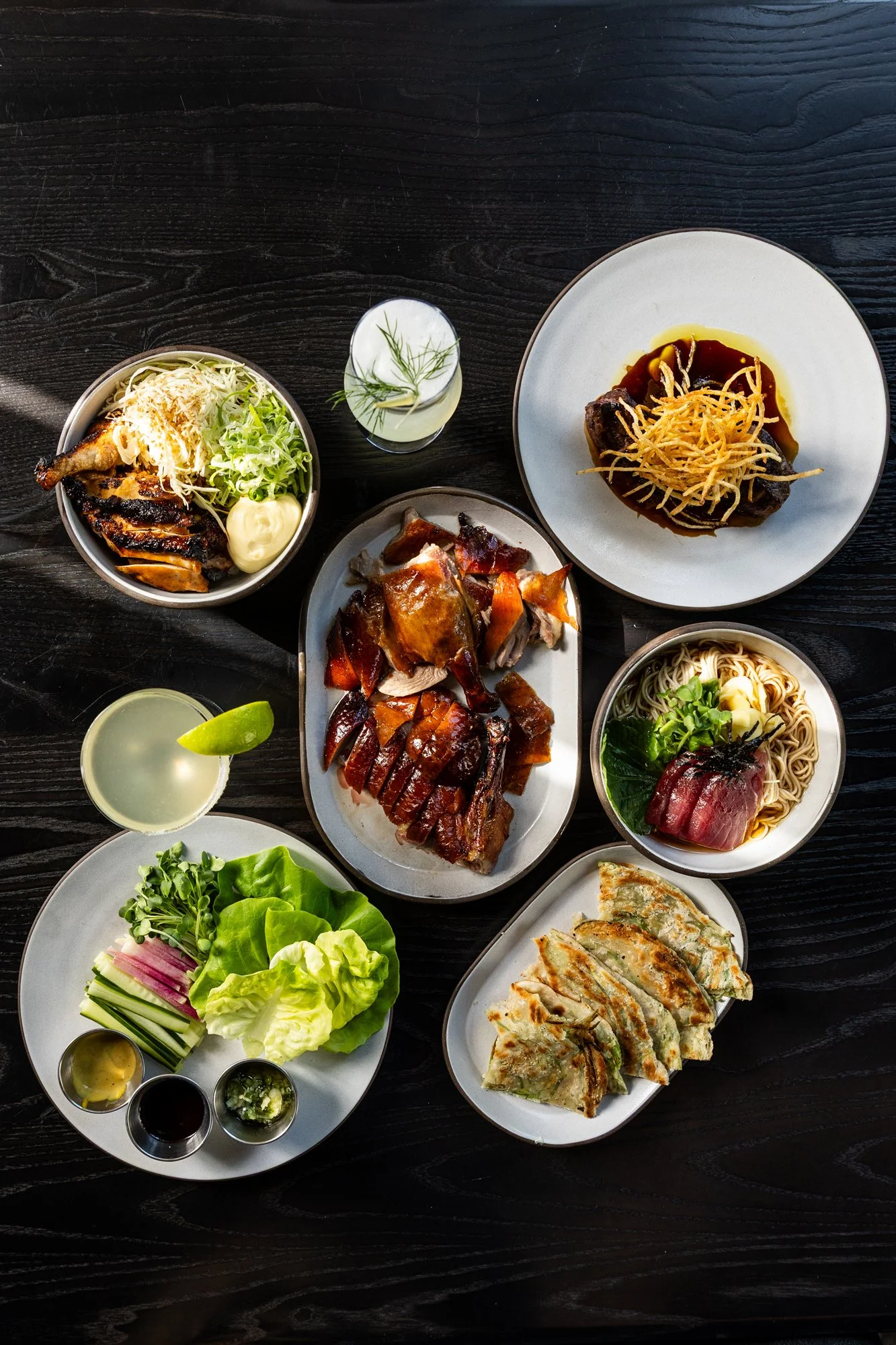 Assorted Asian dishes on a dark wooden table, including grilled chicken, roasted pork, noodles with beef, spring rolls, and various vegetables with dipping sauces, along with beverages.