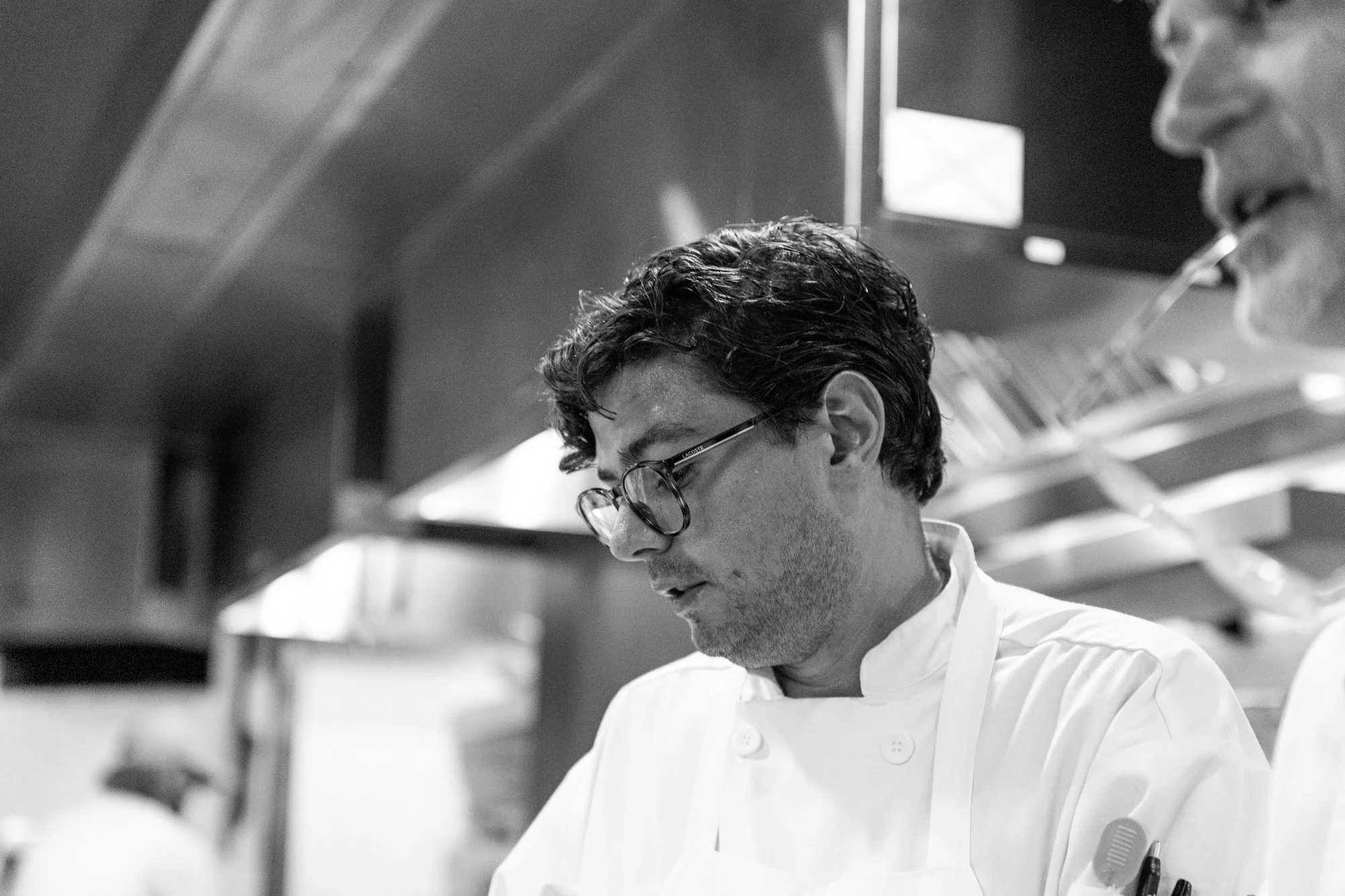 A male chef with glasses and dark, curly hair, wearing a white chef's coat, is focused on cooking in a professional kitchen. Hot andHot Fish Club Restaurant in Birmingham, Alabama.