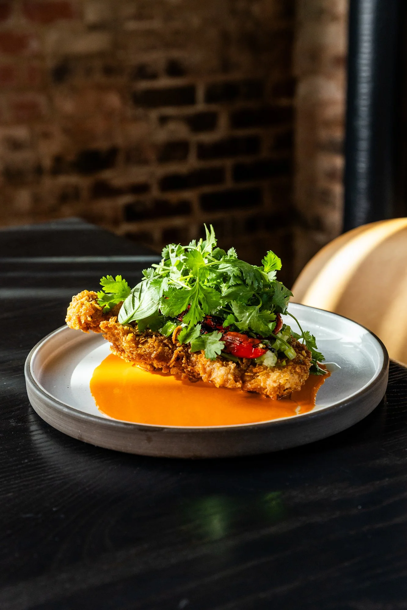 Fried fish fillet topped with fresh greens and herbs on a white plate with orange sauce, set on a dark table in front of a brick wall.
