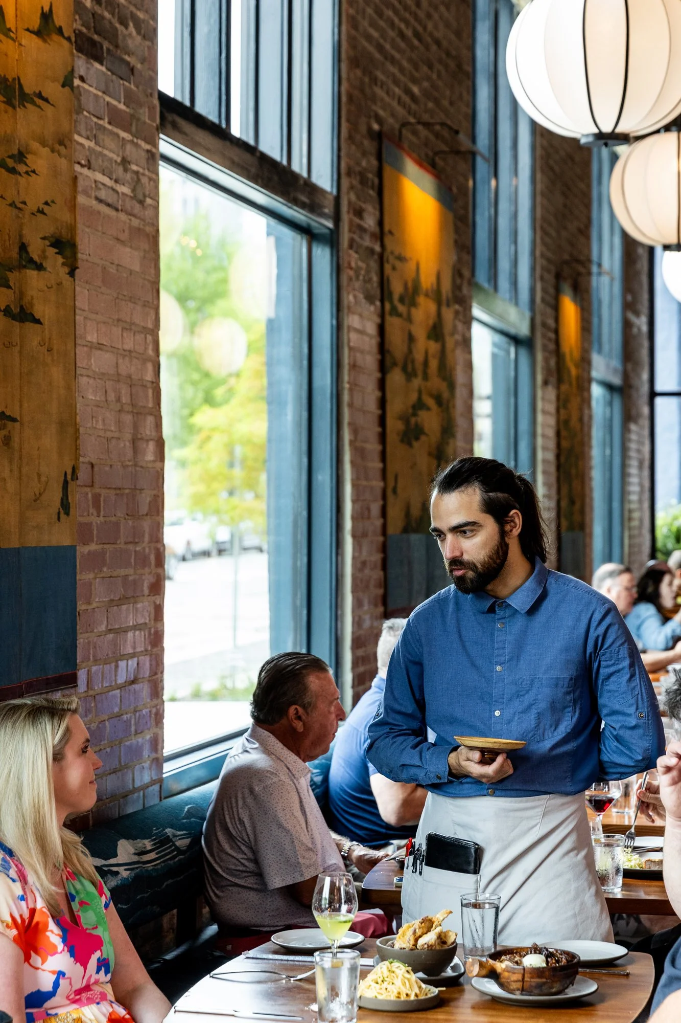 A restaurant scene with a waiter in a blue shirt serving a table where a woman with blonde hair and a colorful dress, and an older man with a gray shirt are seated. The table has various dishes, glasses, and a bowl of fried food.