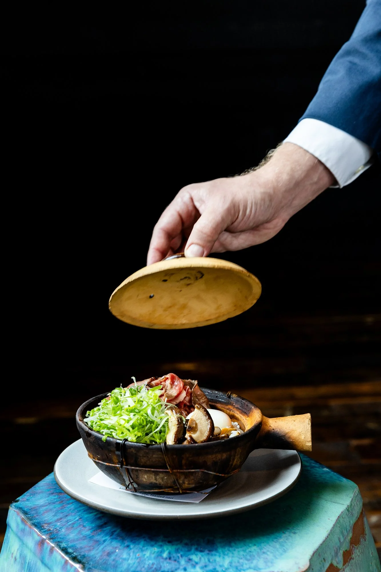 A chef garnishing a bowl of ramen with a wooden lid in a dimly lit setting.