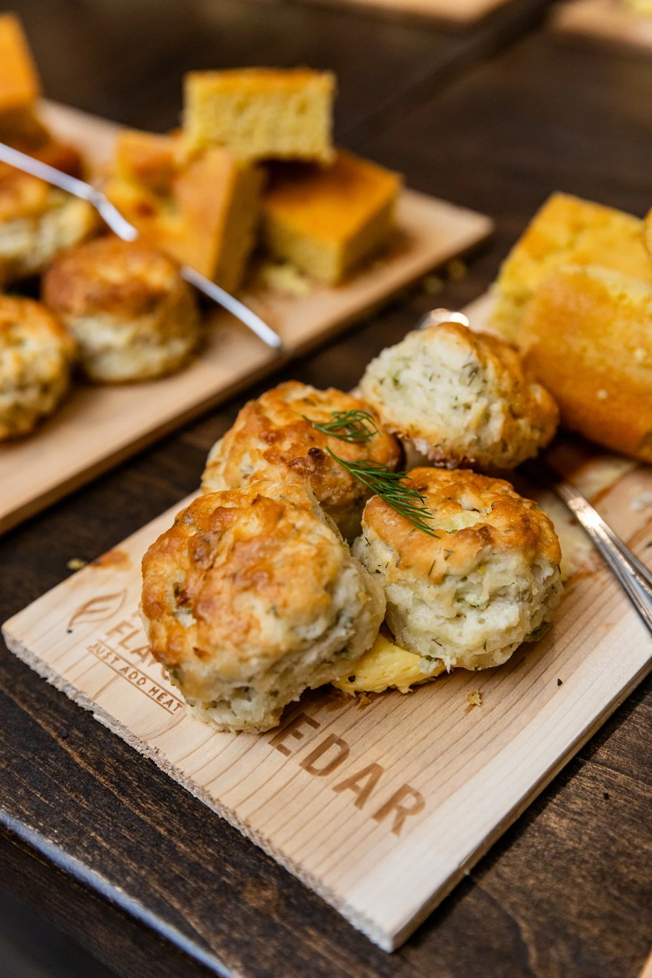 Close-up of four baked chicken meatballs garnished with a small sprig of fresh herb, served on a wooden cutting board. In the background, slices of cornbread and other baked items are visible on another board. Southbound Food Festival in Birmingham, 