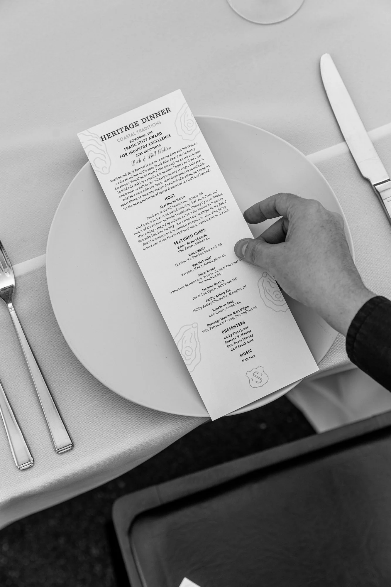 A person holding a program or menu at a formal event, with a white plate, knife, and fork on a table covered with a cloth. The event appears to be a heritage dinner. Southbound Food Festival in Birmingham, Alabama.