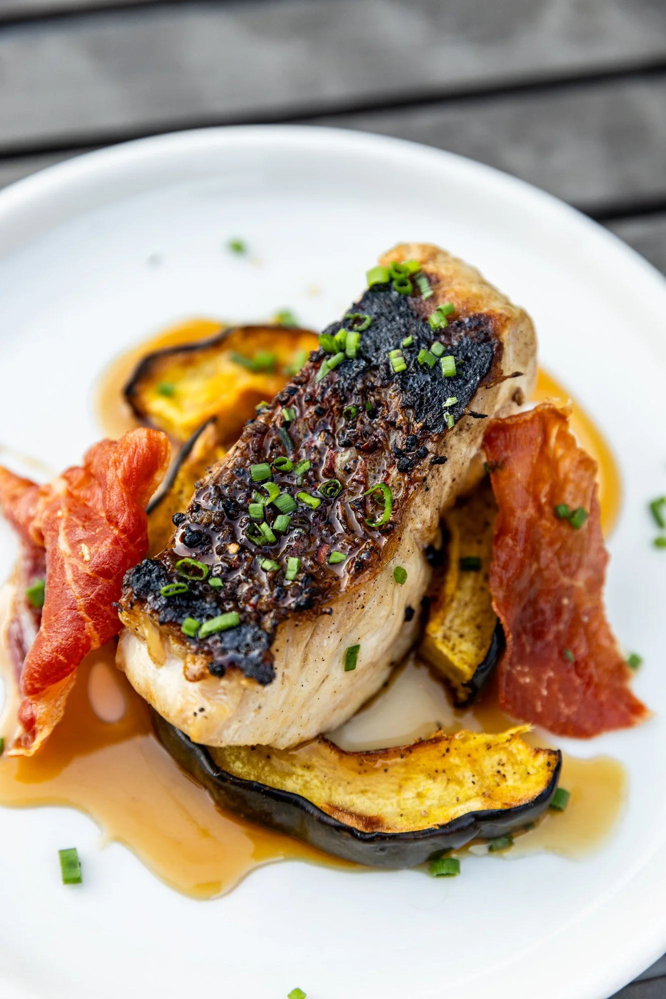 A plated dish featuring a piece of grilled fish with crispy skin topped with chopped green herbs, served with roasted eggplant slices and crispy bacon on a white plate. Ovenbird Restaurant in Birmingham, Alabama.