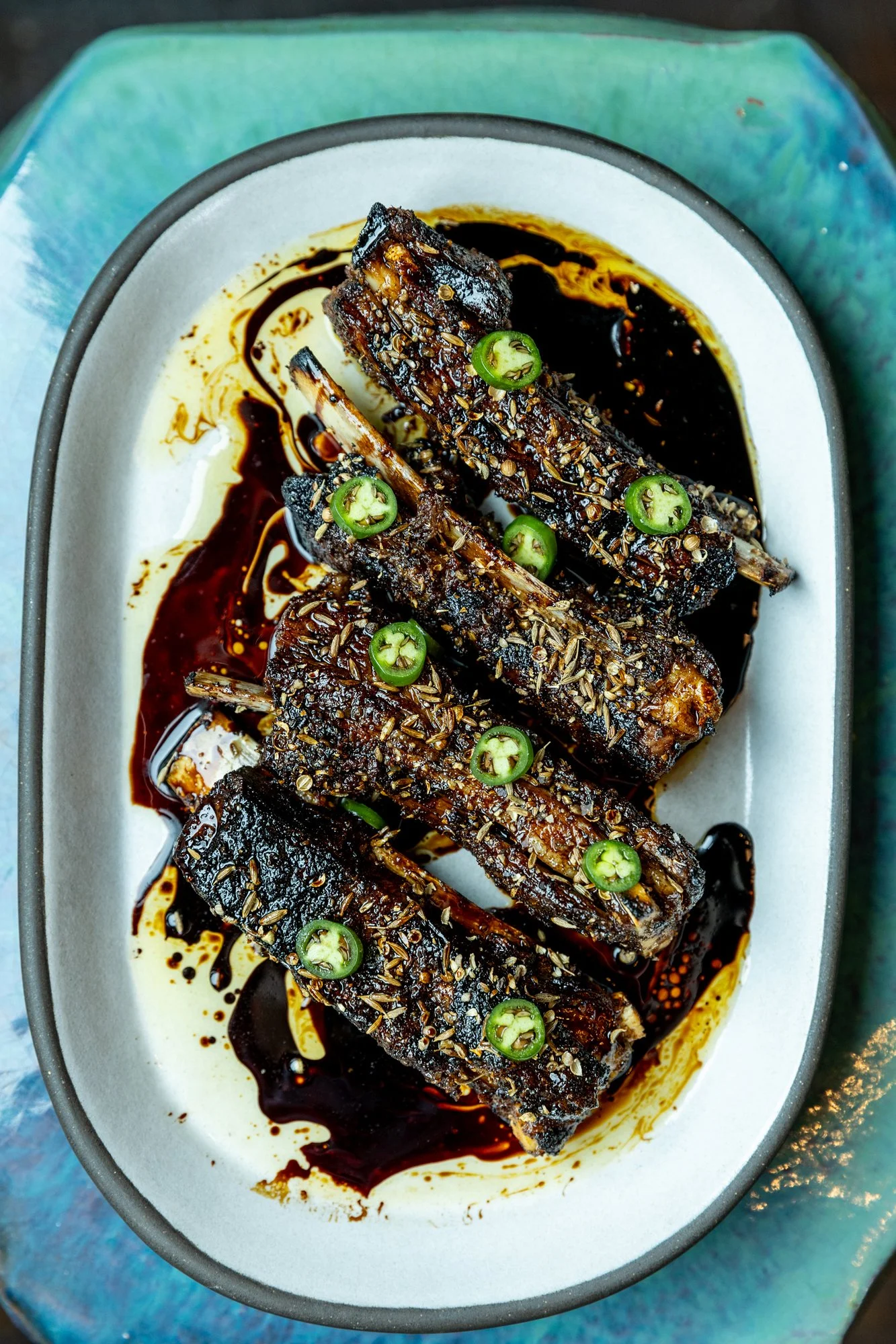 Plate of glazed pork ribs garnished with sliced green chili peppers, sesame seeds, and drizzled with dark barbecue sauce.