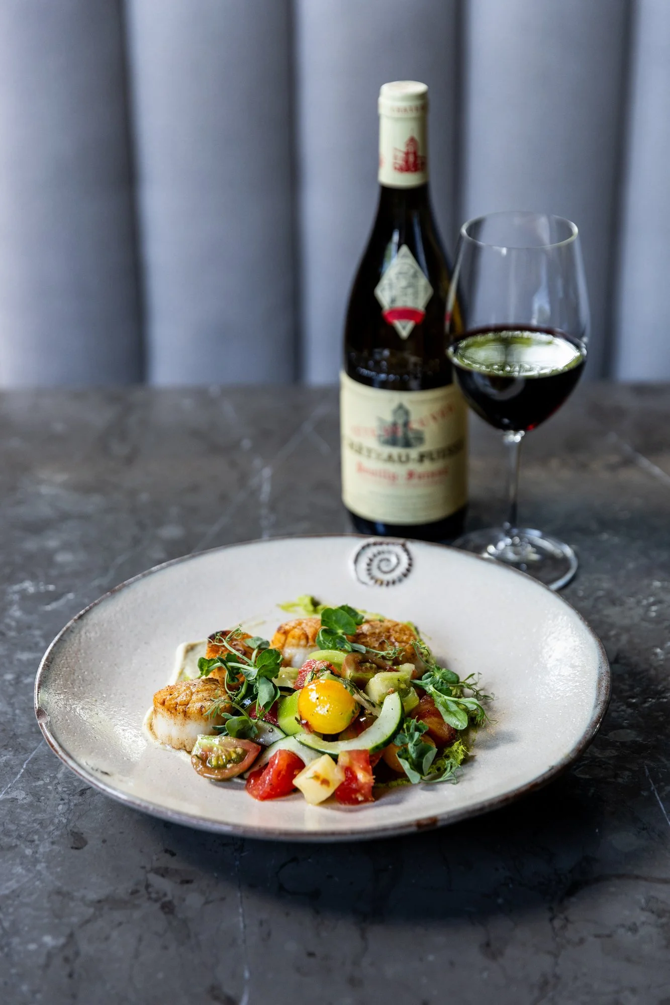 A plate of gourmet salad with cherry tomatoes, cucumber, greens, and a yellow edible flower, with a glass of red wine and a bottle of Beaujolais wine on a dark marble table. Hot and Hot Fish Club Restaurant in Birmingham, Alabama.