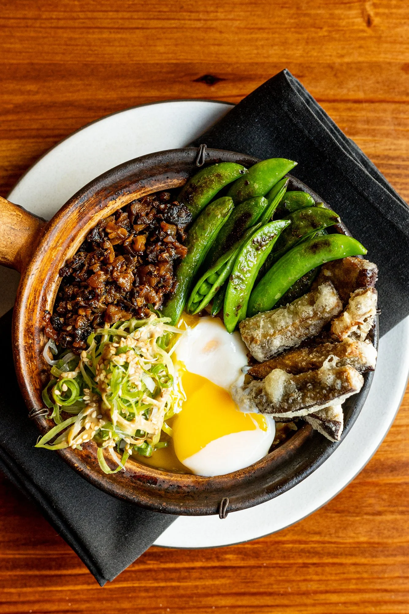 Clay bowl with fried vegetables, snap peas, soft-boiled egg, chopped green onions, and bean paste, on a black napkin over a wooden table.