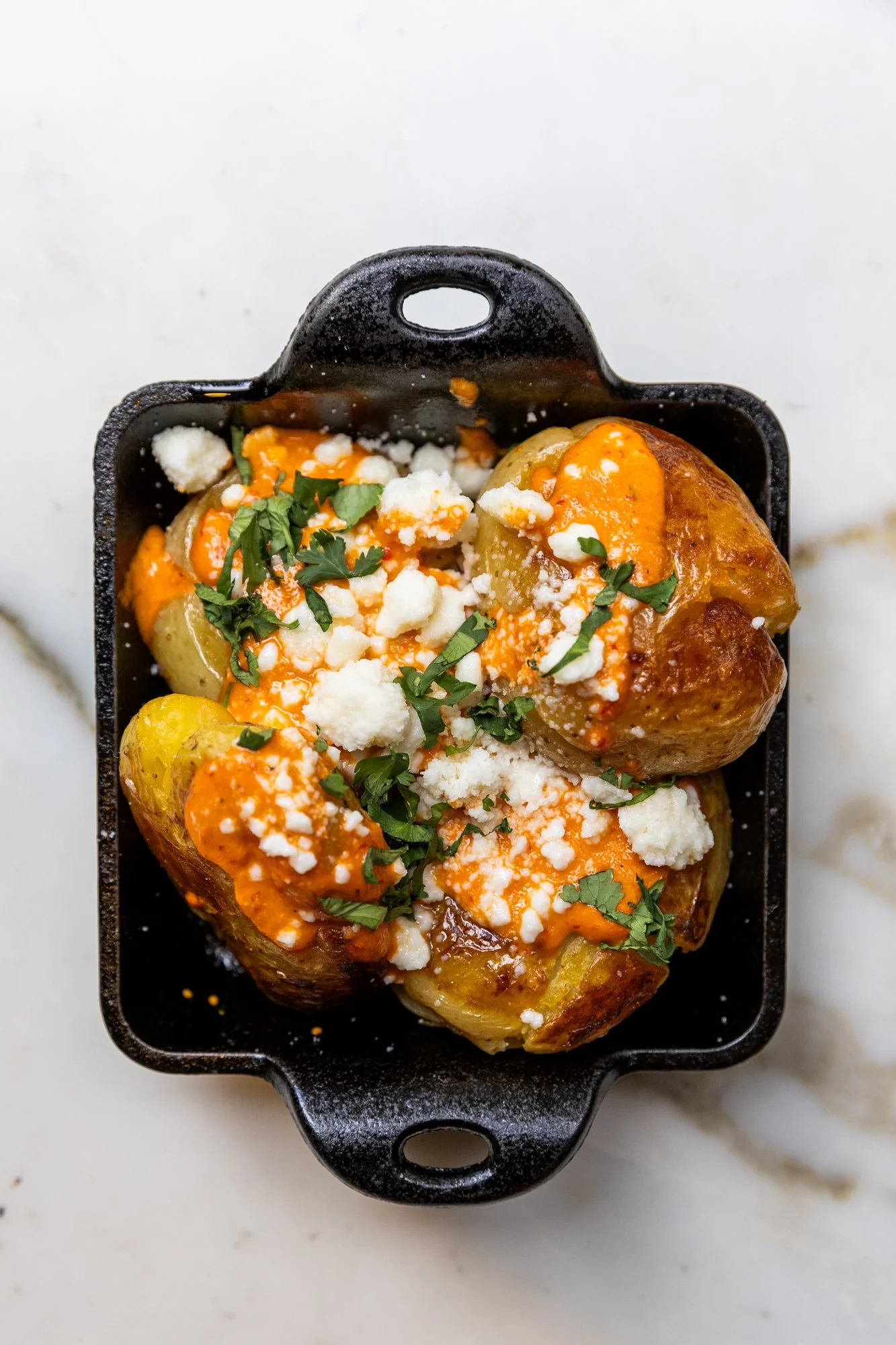 Baked potatoes topped with cheese, chopped herbs, and sauce in a black baking dish Ovenbird Restaurant in Birmingham, Alabama.