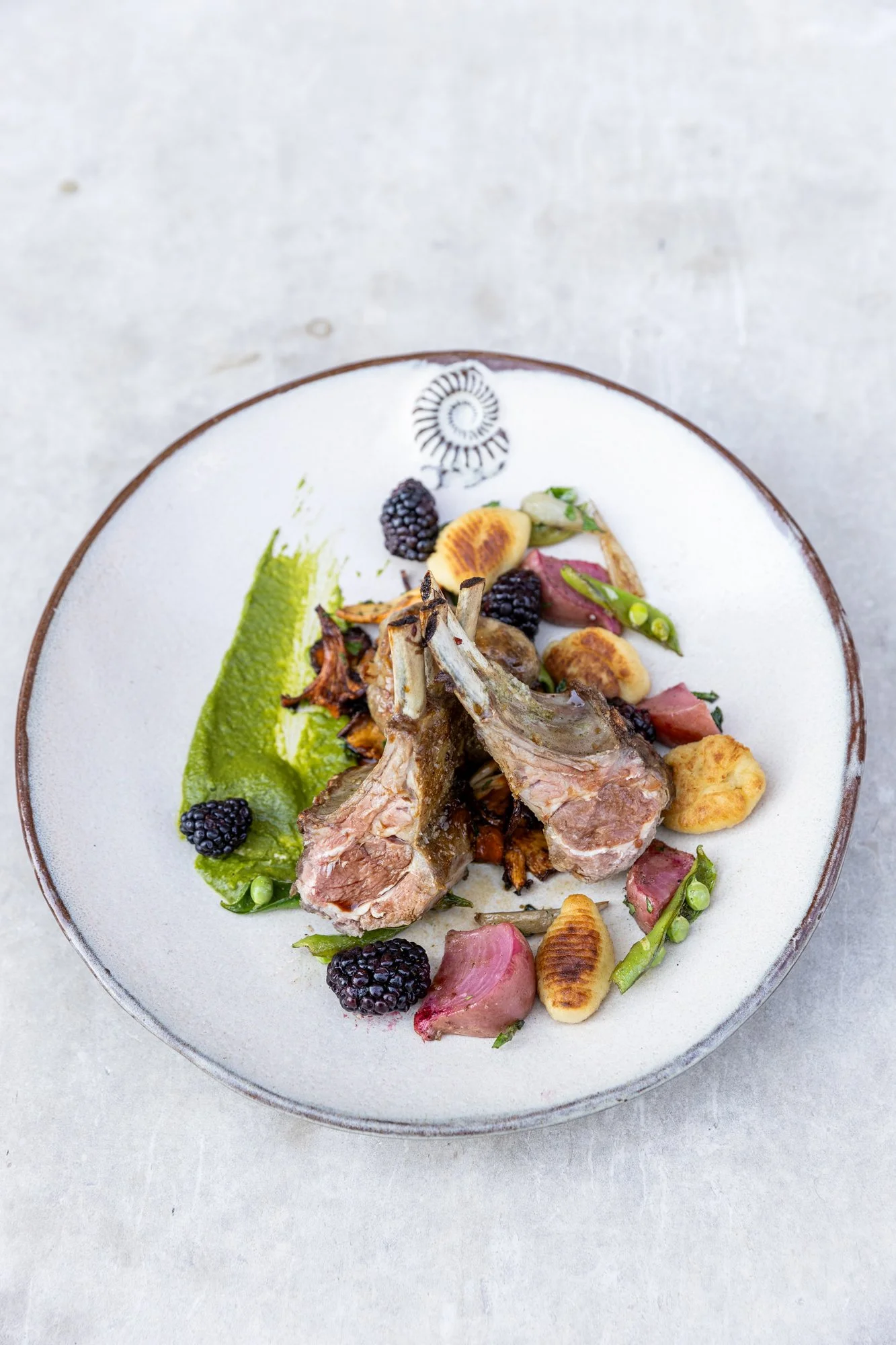 A plated dish featuring grilled lamb chops, blackberries, sliced radishes, green peas, and a green sauce on a white ceramic plate. Hot and Hot Fish Club Restaurant in Birmingham, Alabama.