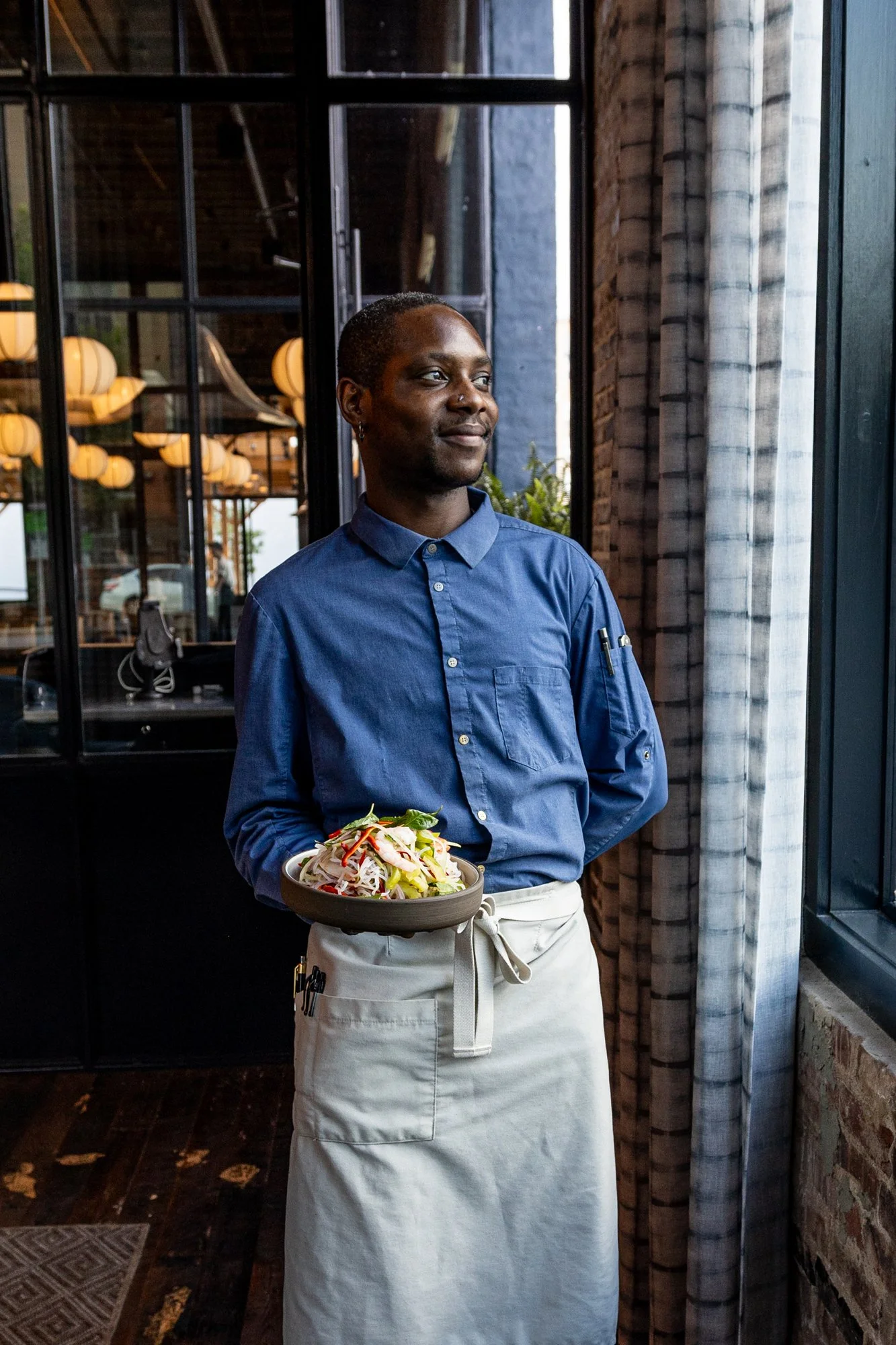 A male chef standing indoors near a window, holding a bowl of salad, wearing a blue chef's jacket and an apron.
