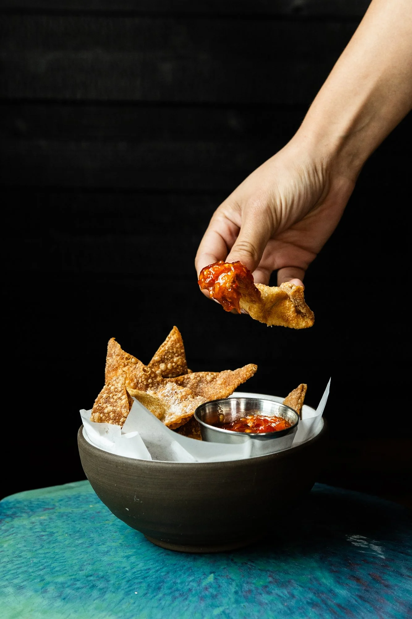 A hand holding a crispy fried chicken wing dipped in sauce over a bowl of fried tortilla chips and salsa.
