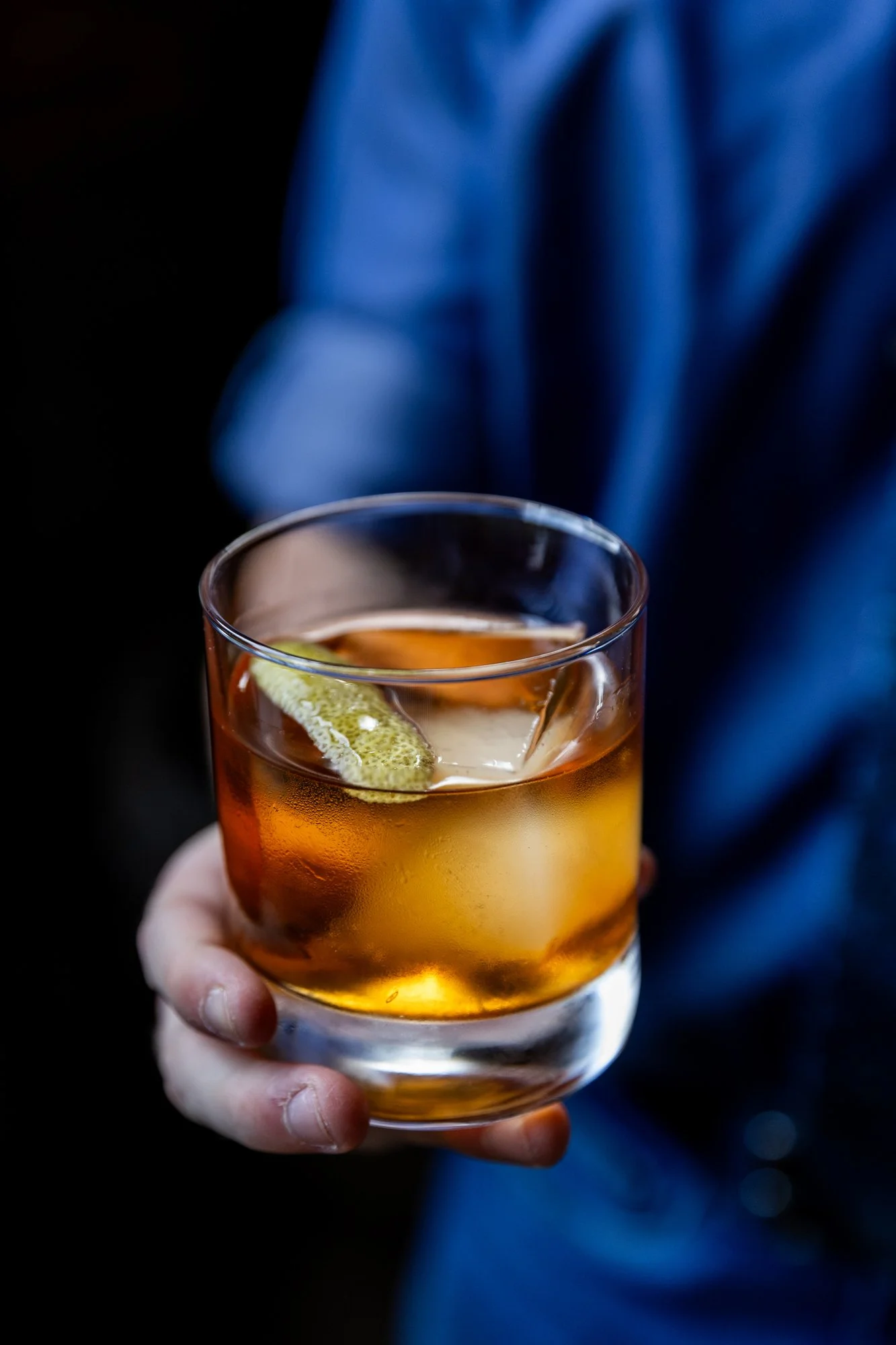 Person holding a glass of amber-colored cocktail with a lime wedge and ice, wearing a blue shirt. Hot and Hot Fish Club Restaurant in Birmingham, Alabama.