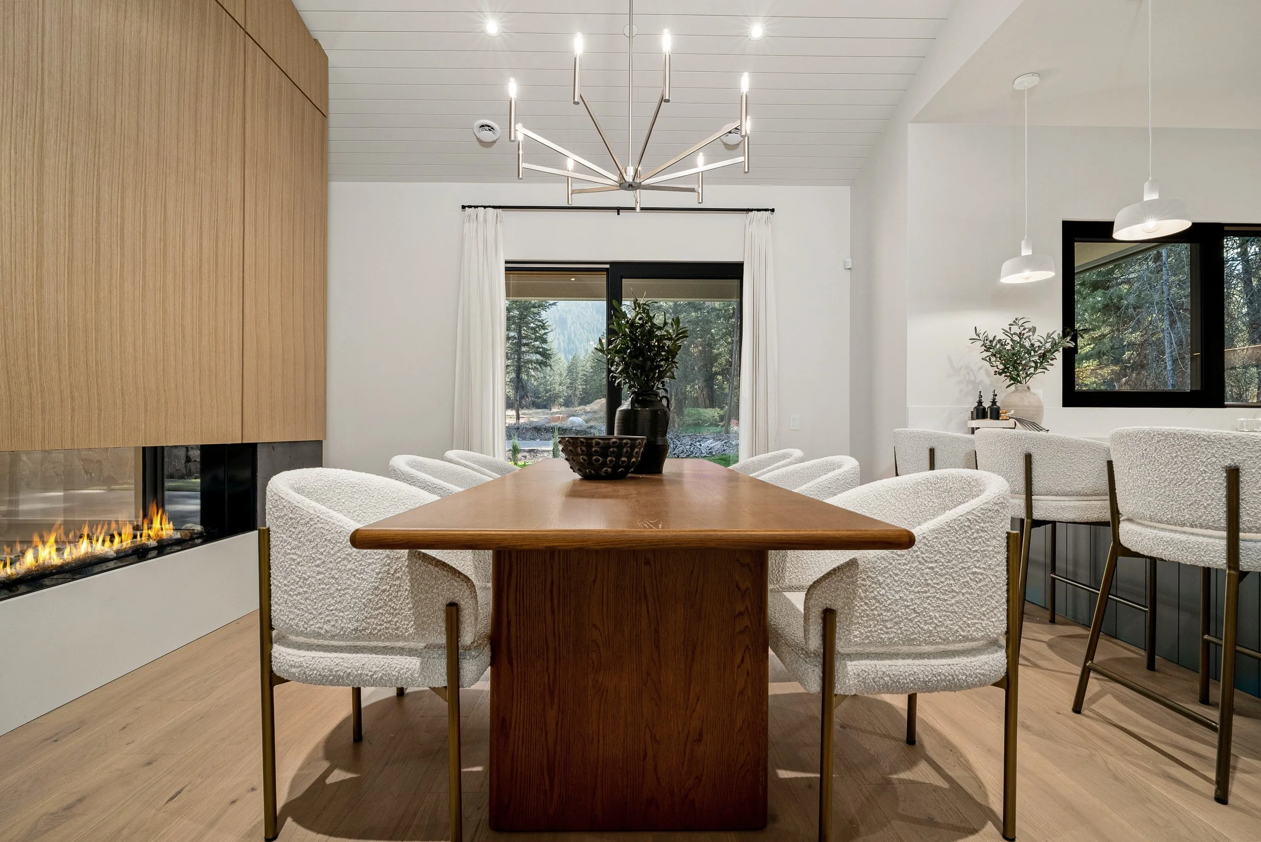 Dining at Woodside Showhome — wood table, bouclé chairs, and the fireplace wall opening to forest views