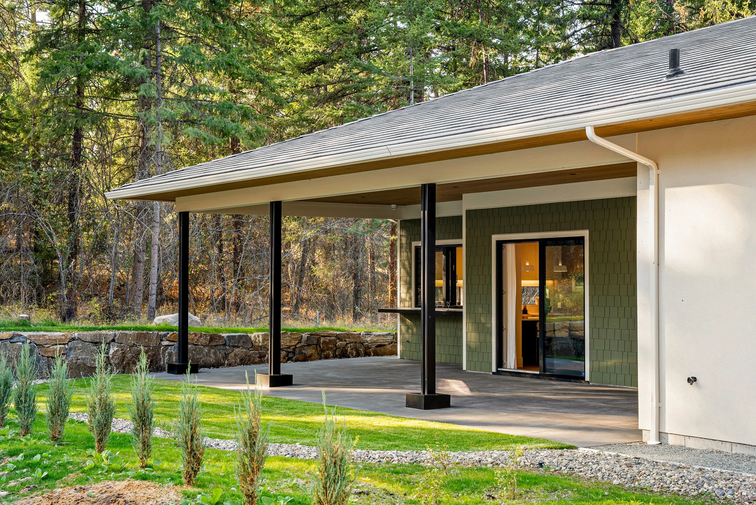 Covered patio at Woodside Showhome with black posts, sage shingle siding and a lawn at the forest edge