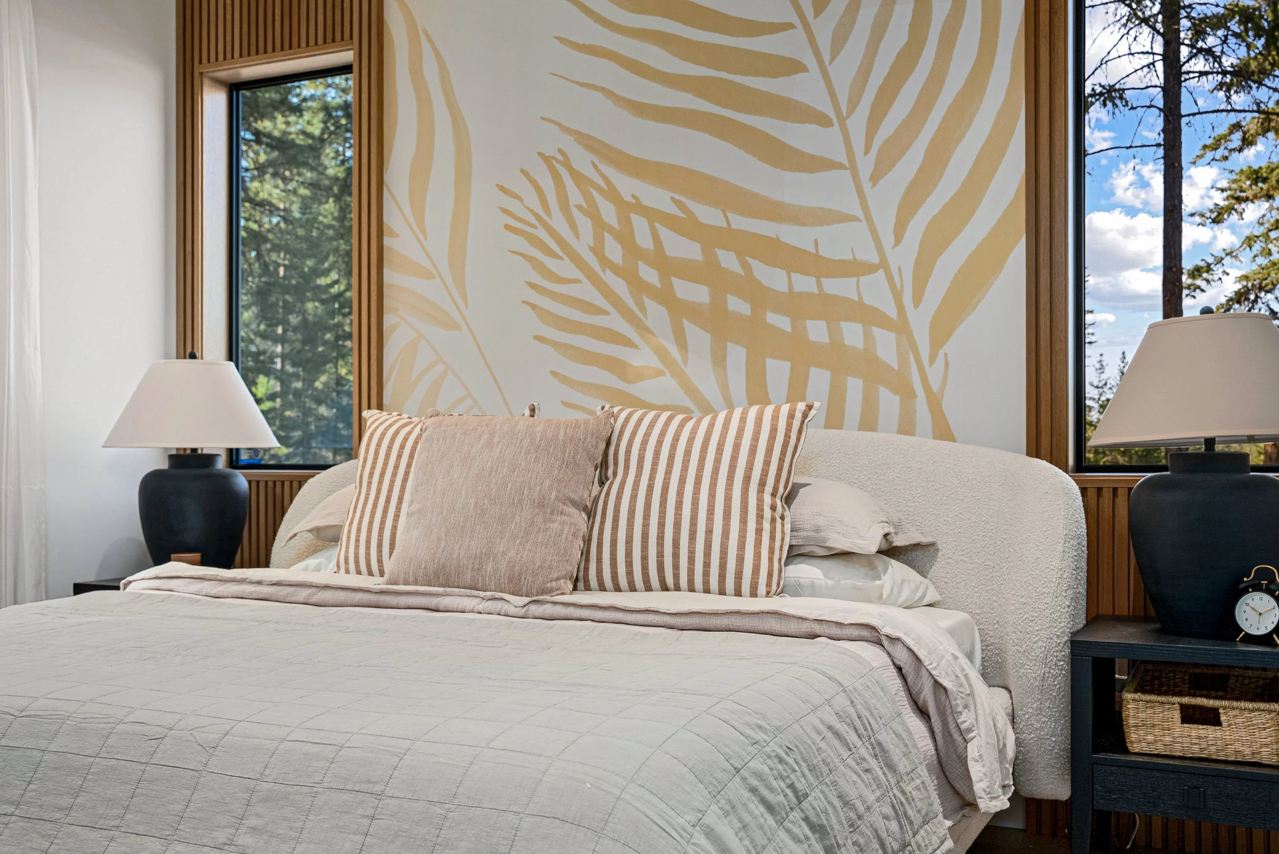 Close view of a Woodside Showhome bed with a bouclé headboard, striped pillows and black lamps against the palm-leaf mural