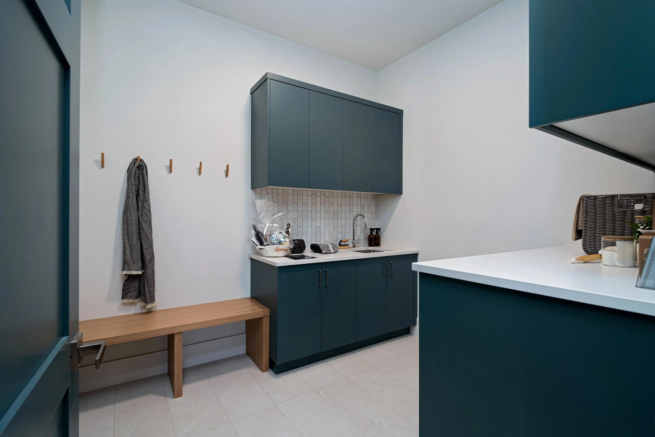 Woodside Showhome mudroom with teal cabinets and sink, a tiled backsplash, wood bench and coat hooks