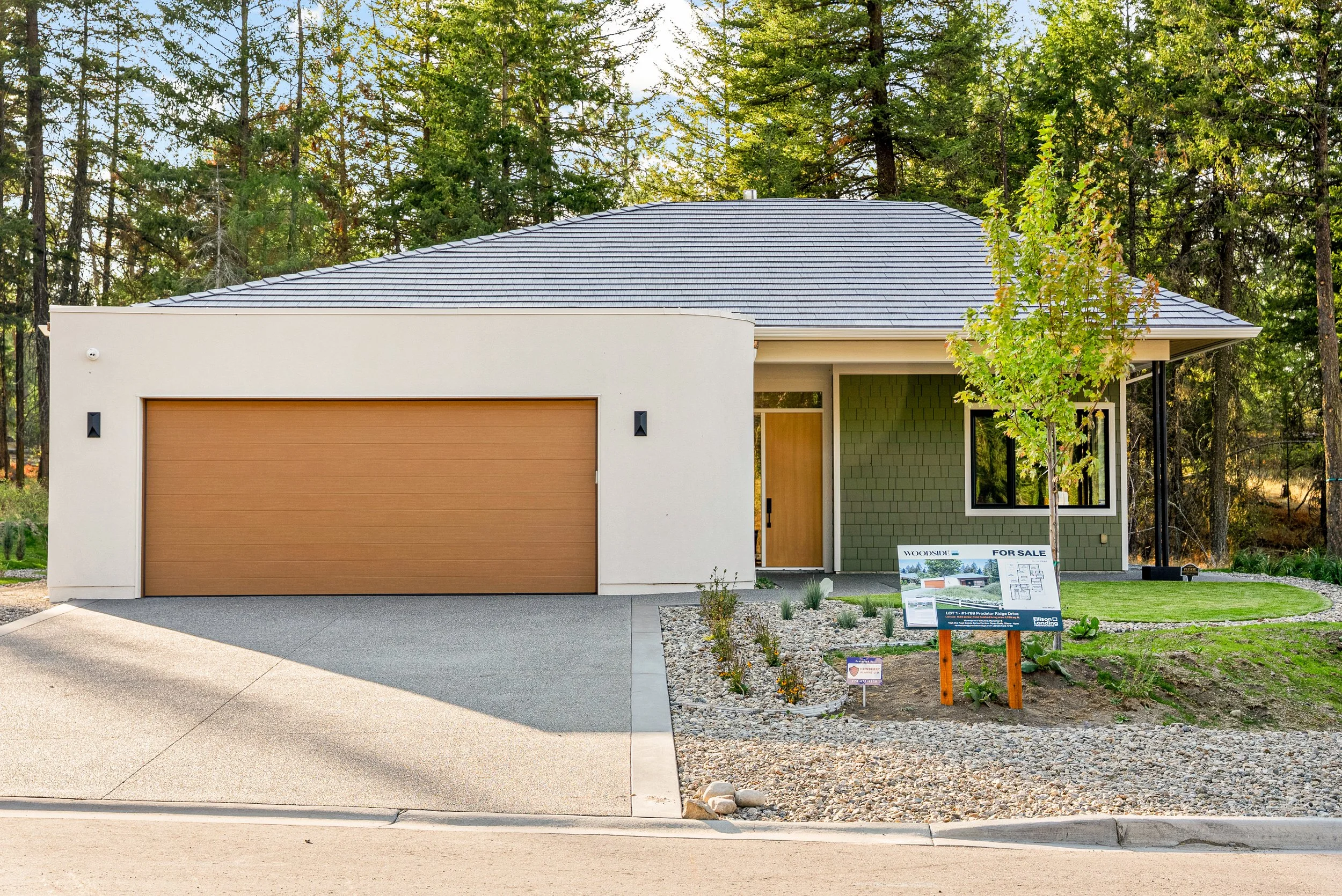 Woodside Showhome exterior at Predator Ridge with a white stucco garage, sage shingle siding and tall pines