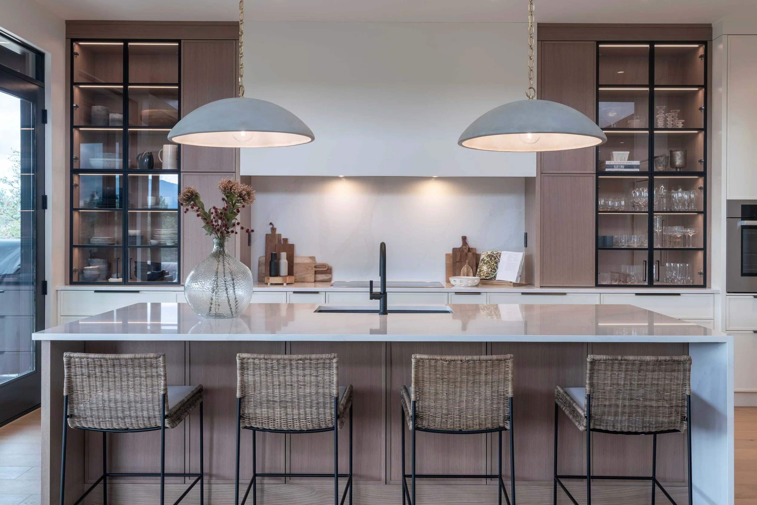 Ridge Light House kitchen with a white island, woven counter stools, dome pendants and black-framed glass cabinets