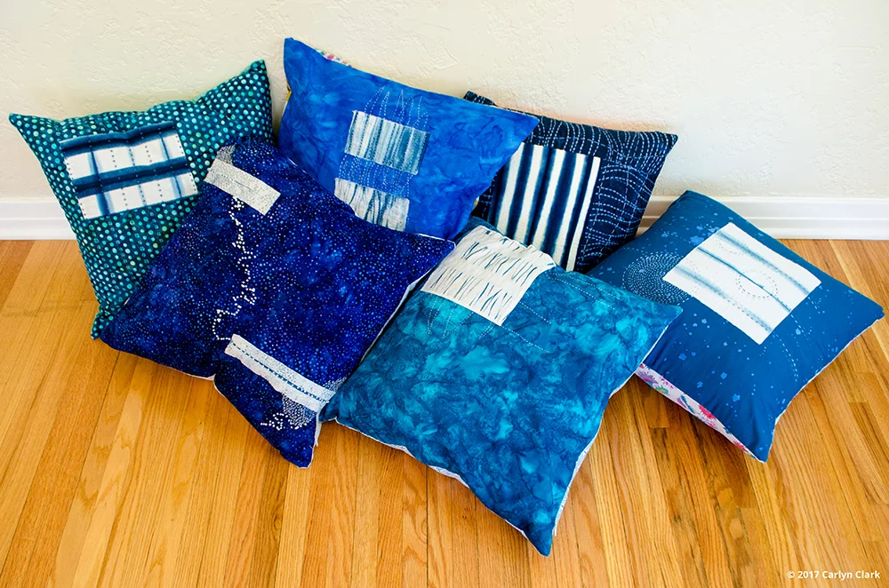 Arrangement of hand-dyed indigo stitched decorative pillows with geometric and sashiko patterns resting on a wooden floor.