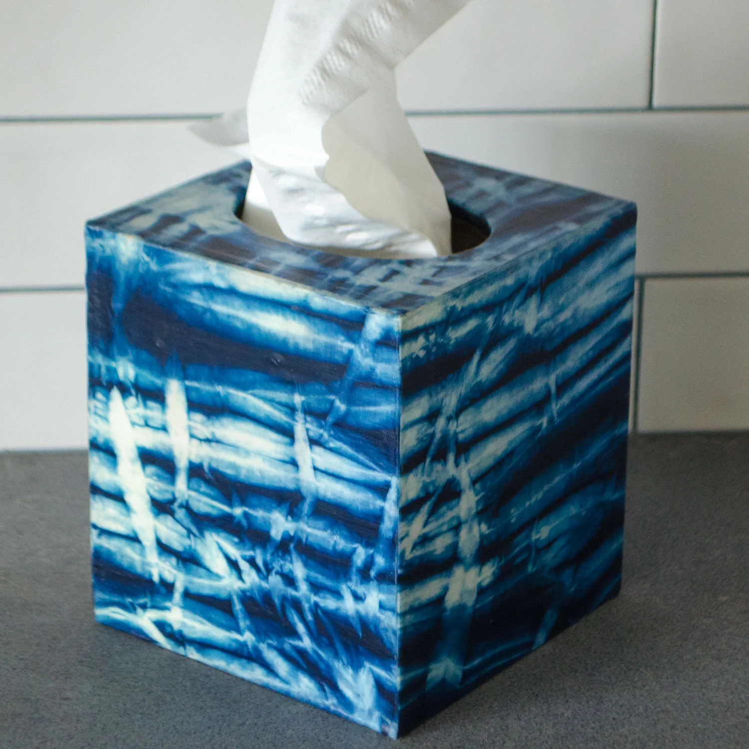 Wooden tissue box cover wrapped in blue and white arashi shibori fabric with a tissue pulling out of the top, sitting on a dark gray countertop.