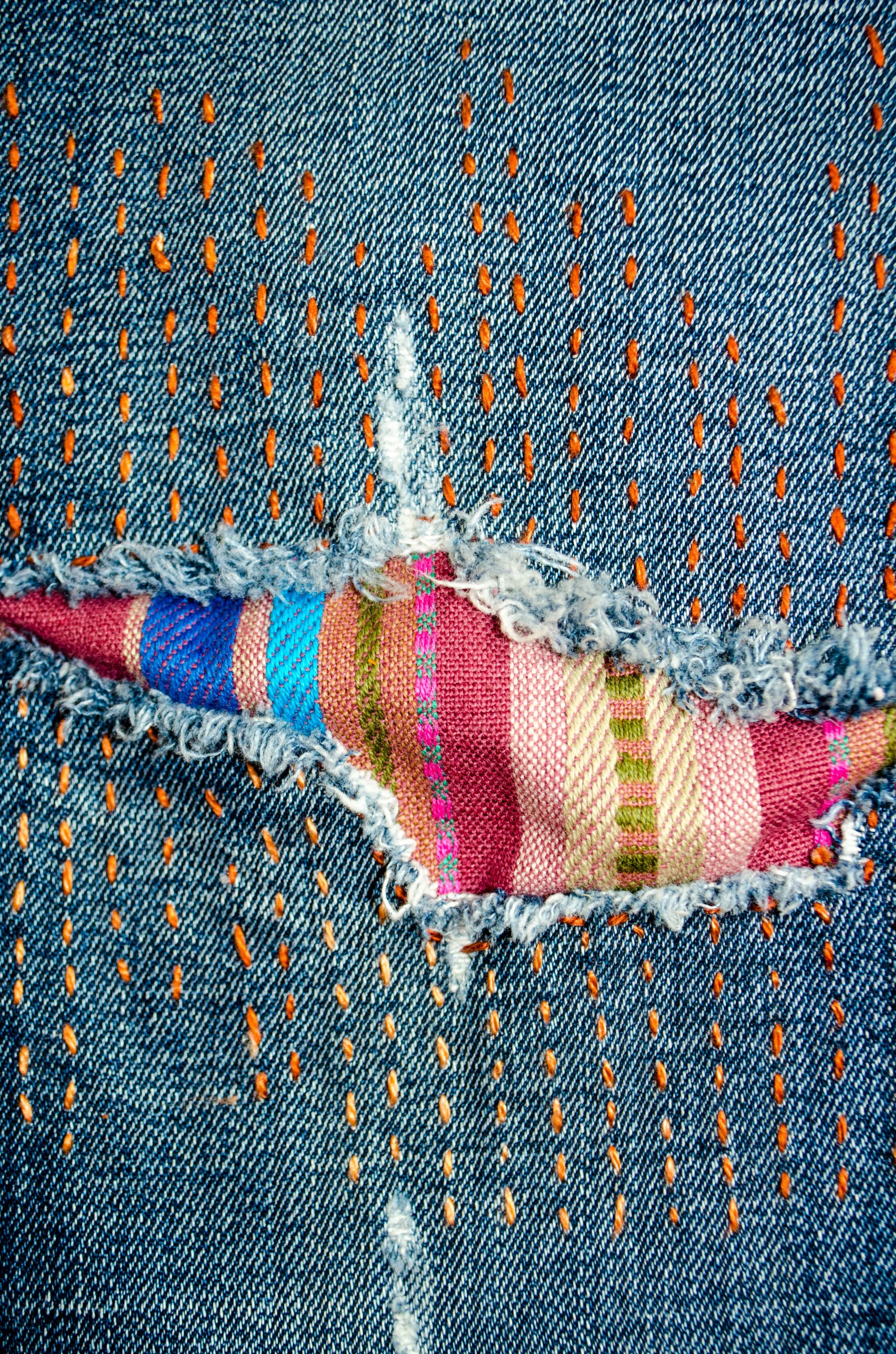 Close-up of visible mending on denim jeans, featuring a bright striped fabric patch peeking through a frayed tear with orange sashiko-inspired running stitches.