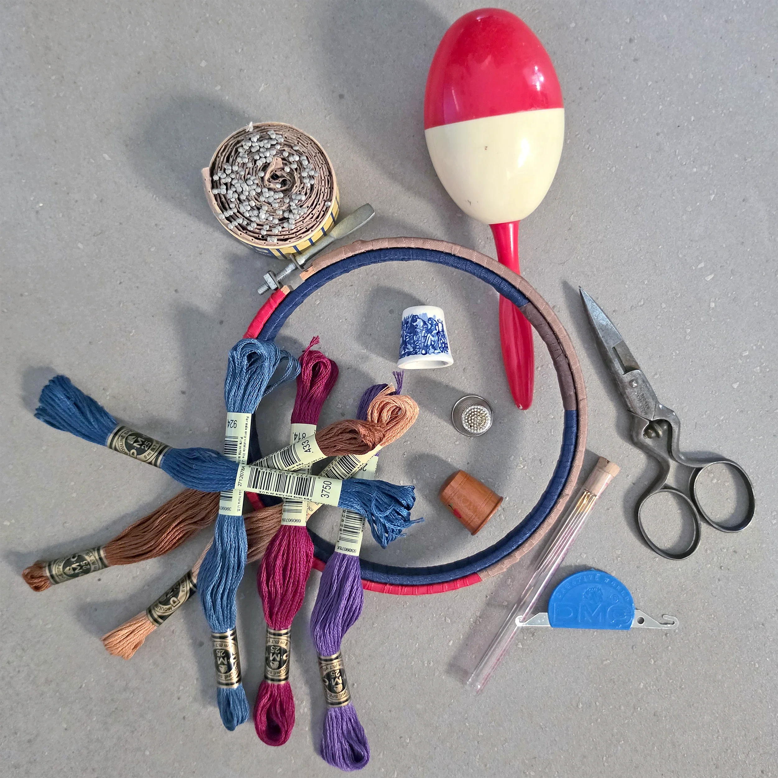 Flat lay of hand embroidery tools on a grey surface: six skeins of DMC floss, a wrapped embroidery hoop, three thimbles, small vintage scissors, a needle case, a roll of pins, a red and cream darning egg, and a blue DMC needle threader.