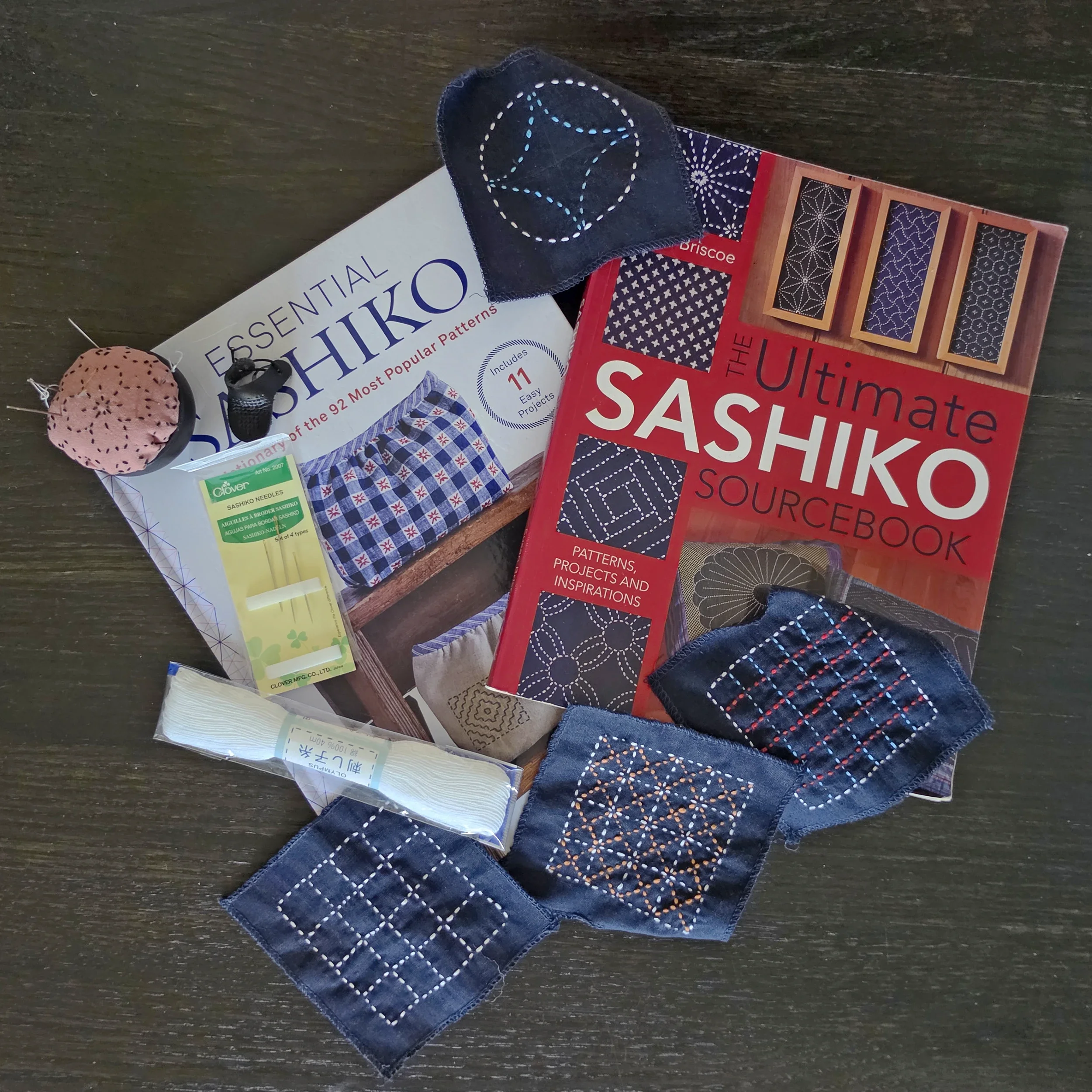 Two sashiko books, The Ultimate Sashiko Sourcebook and Simply Sashiko, on a dark wood surface with navy swatches stitched in white sashiko patterns, a package of needles, and a pincushion.