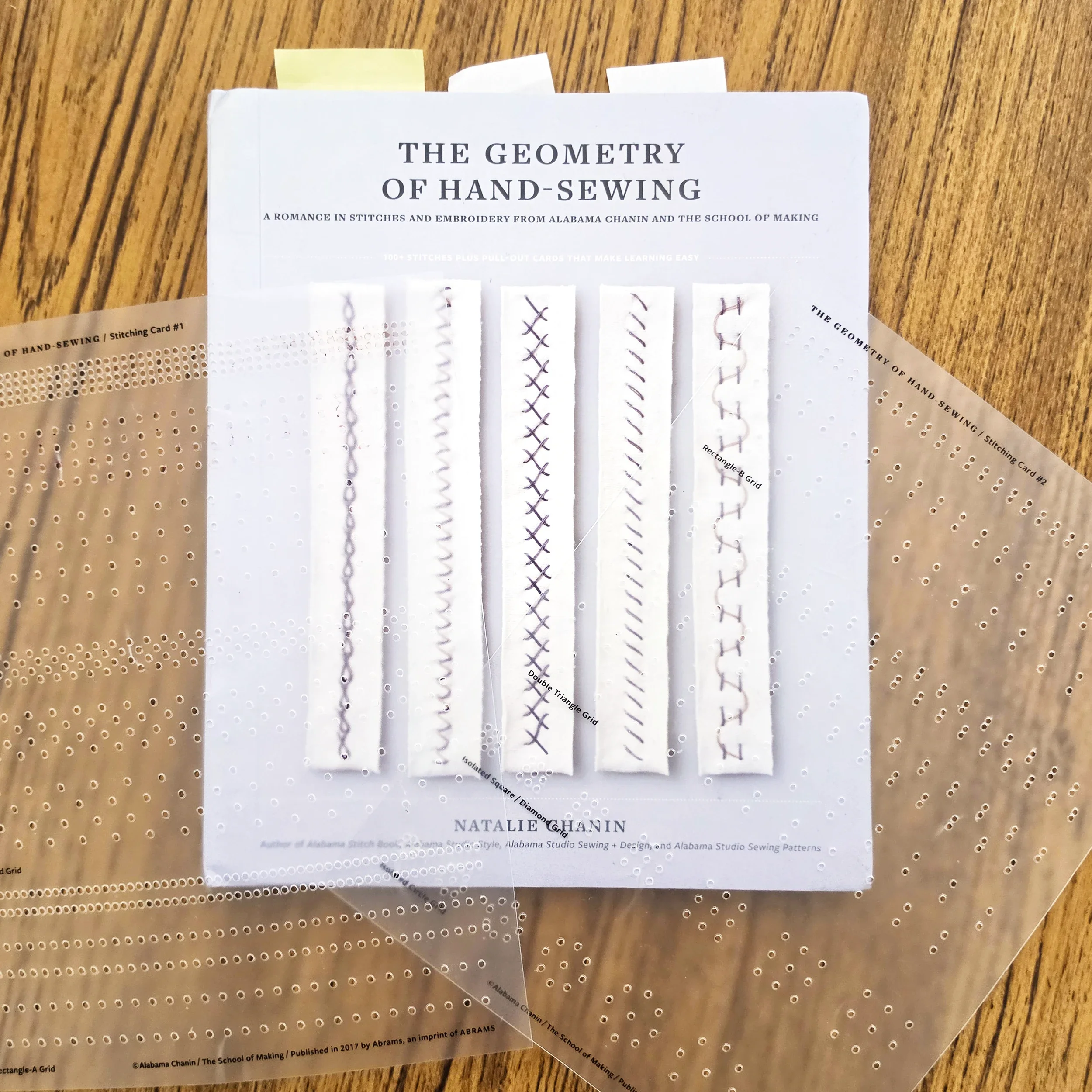 The Geometry of Hand-Sewing by Natalie Chanin, a hand-stitching reference book, shown with its perforated stitching cards on a wooden table