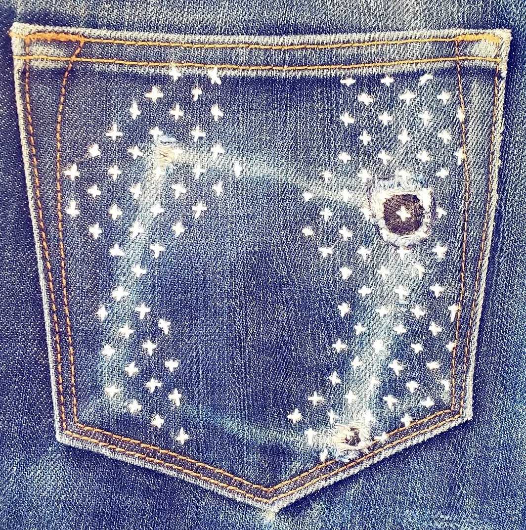 A blue denim back pocket of Railcar Fine Goods jeans featuring intricate white hand stitching inspired by Japanese needlework traditions. Small cross stitches form a geometric pattern across the fabric following the worn wear marks. The design incorp