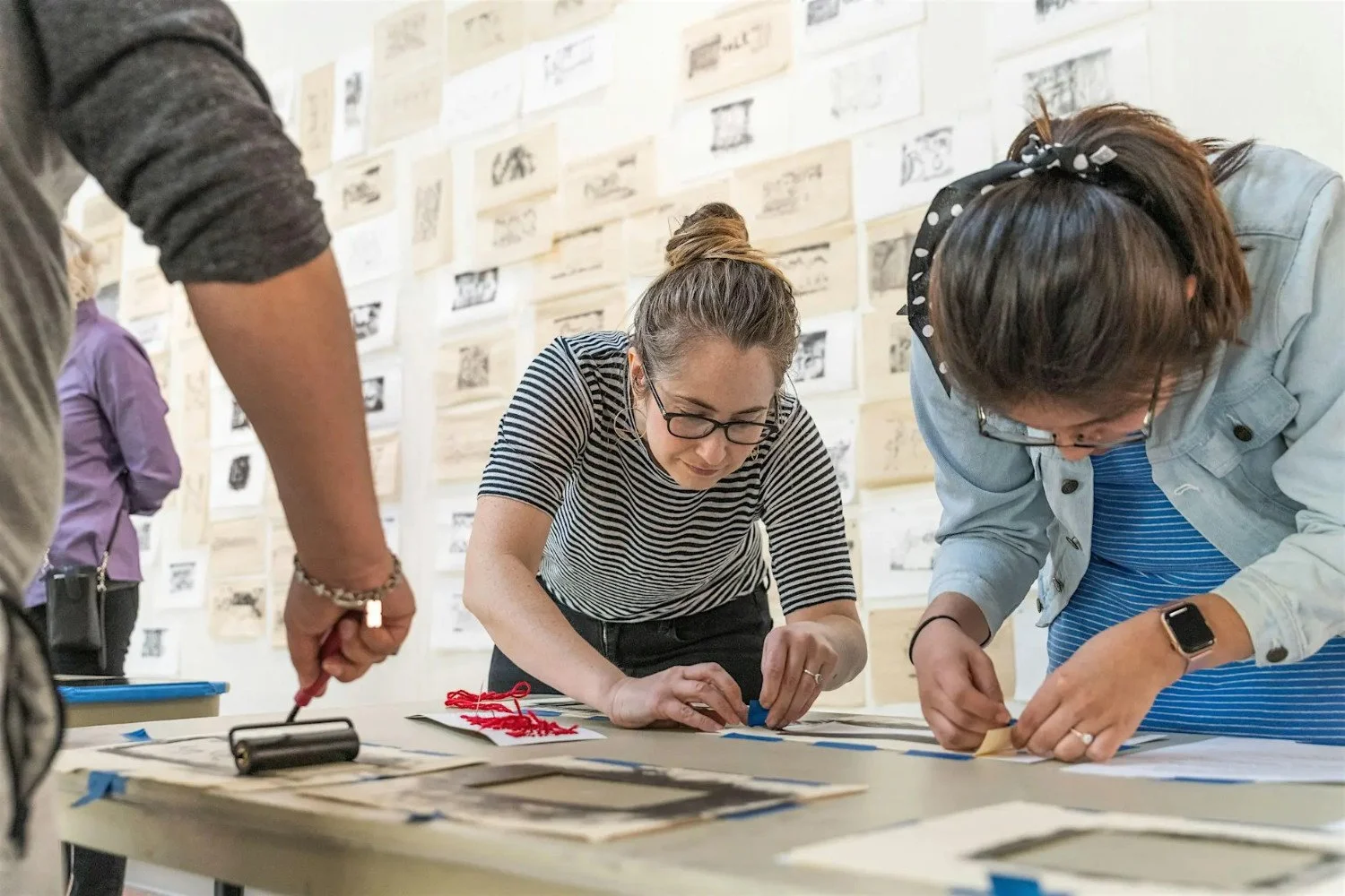People bending over a table, taping or using ink brayers, with prints on the wall behind
