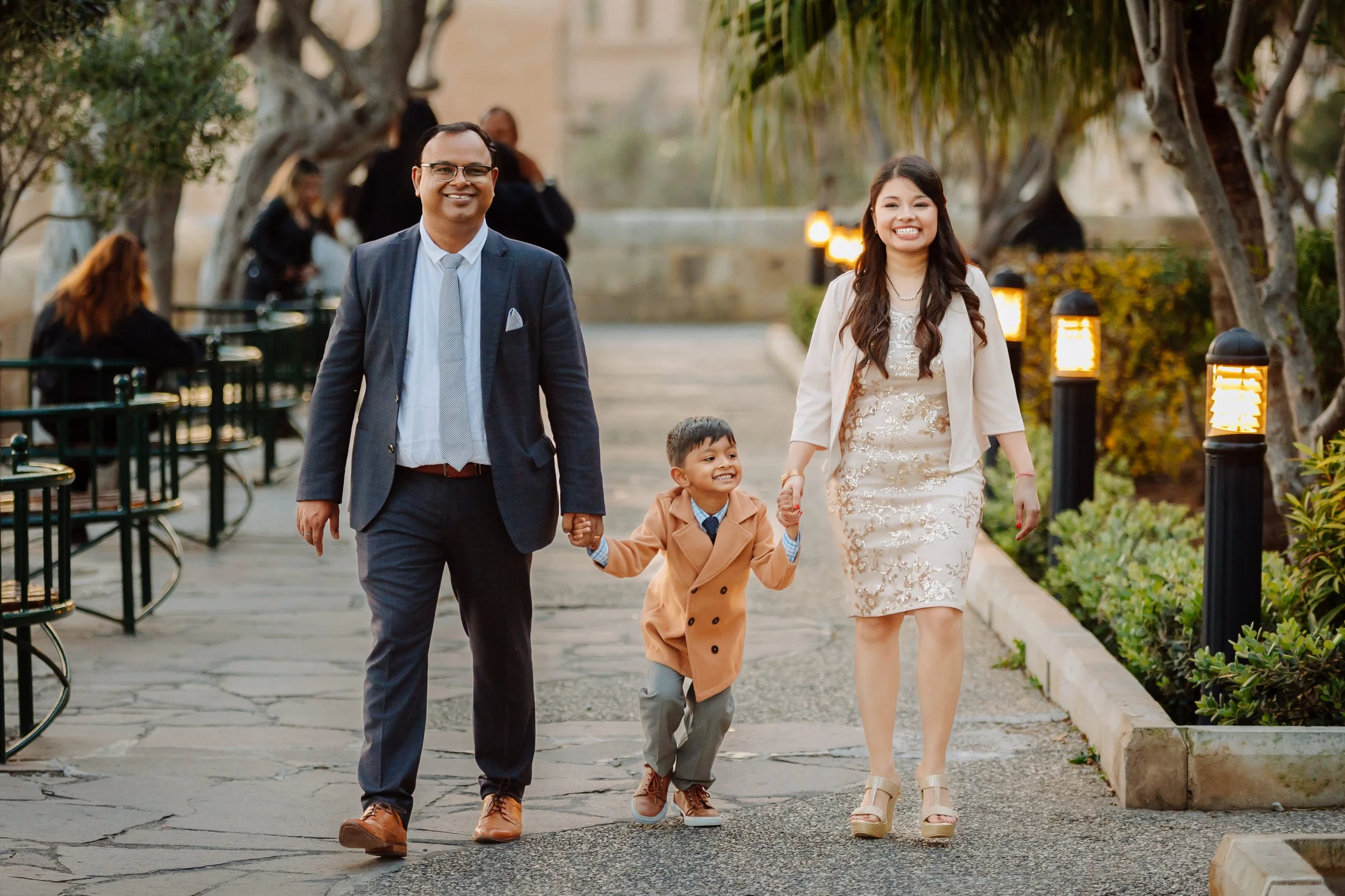 A family of three, including a man in a suit, a woman in a cream dress, and a young boy in a tan coat, walking hand in hand outdoors on a paved pathway lined with trees and lights, smiling and enjoying their time together in a park-like setting.
