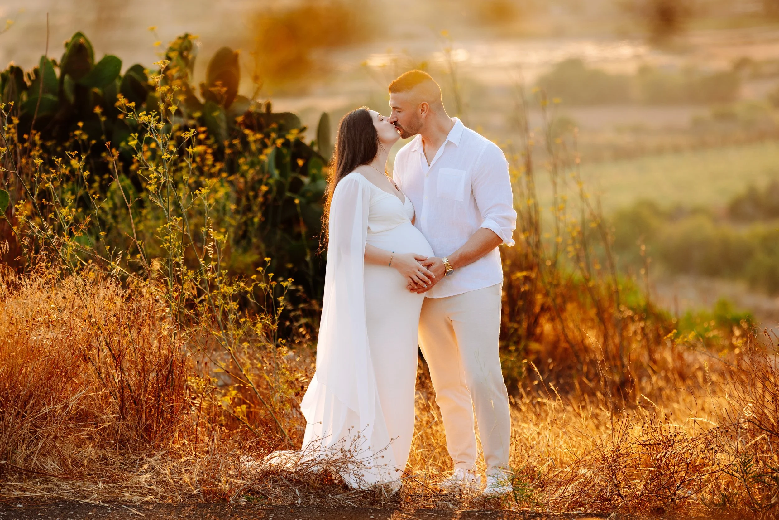 A pregnant woman and a man are sharing a kiss in a field at sunset, both dressed in white.