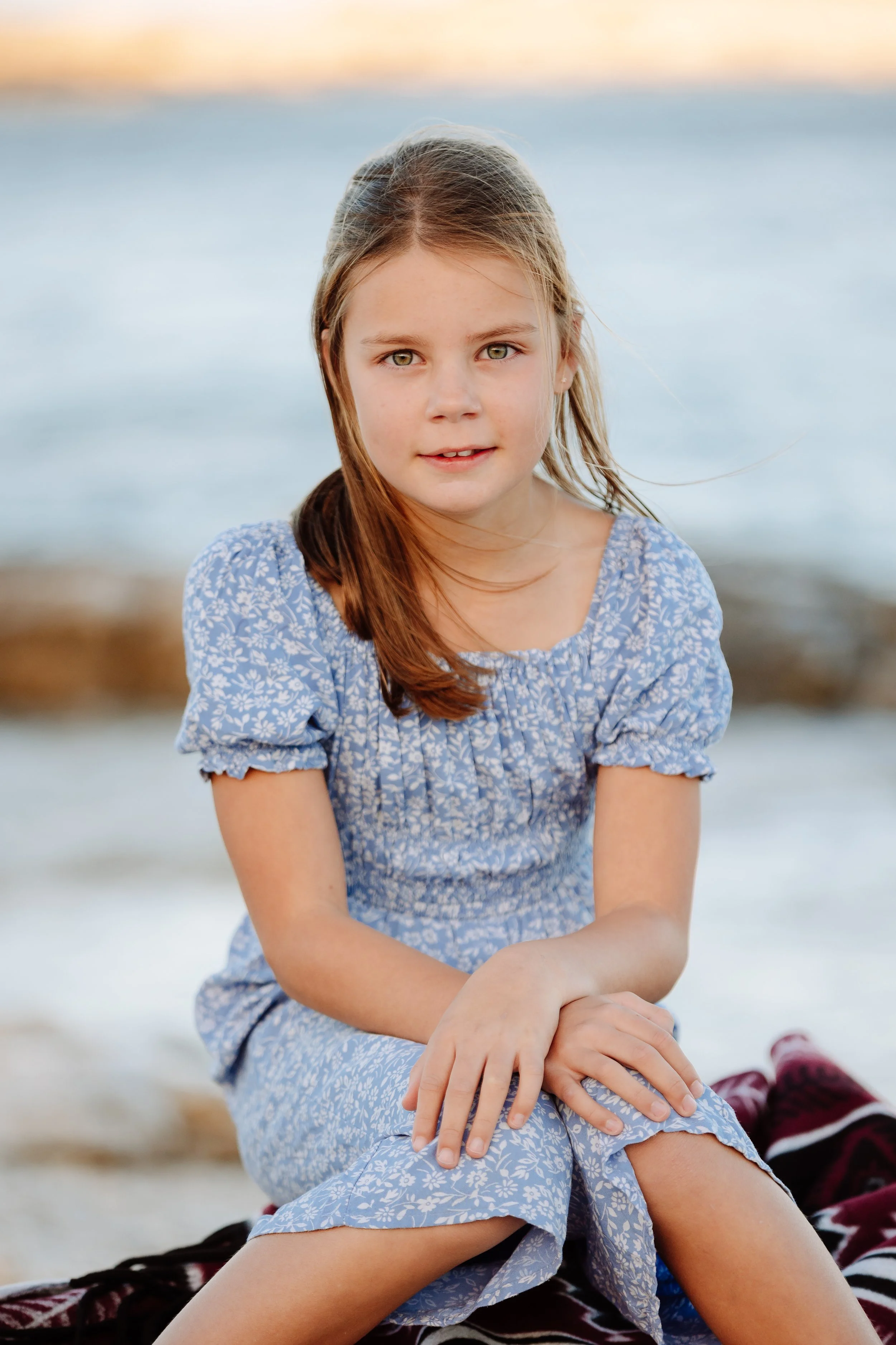 A young girl with long brown hair and green eyes sitting on a beach in a blue floral dress during sunset.