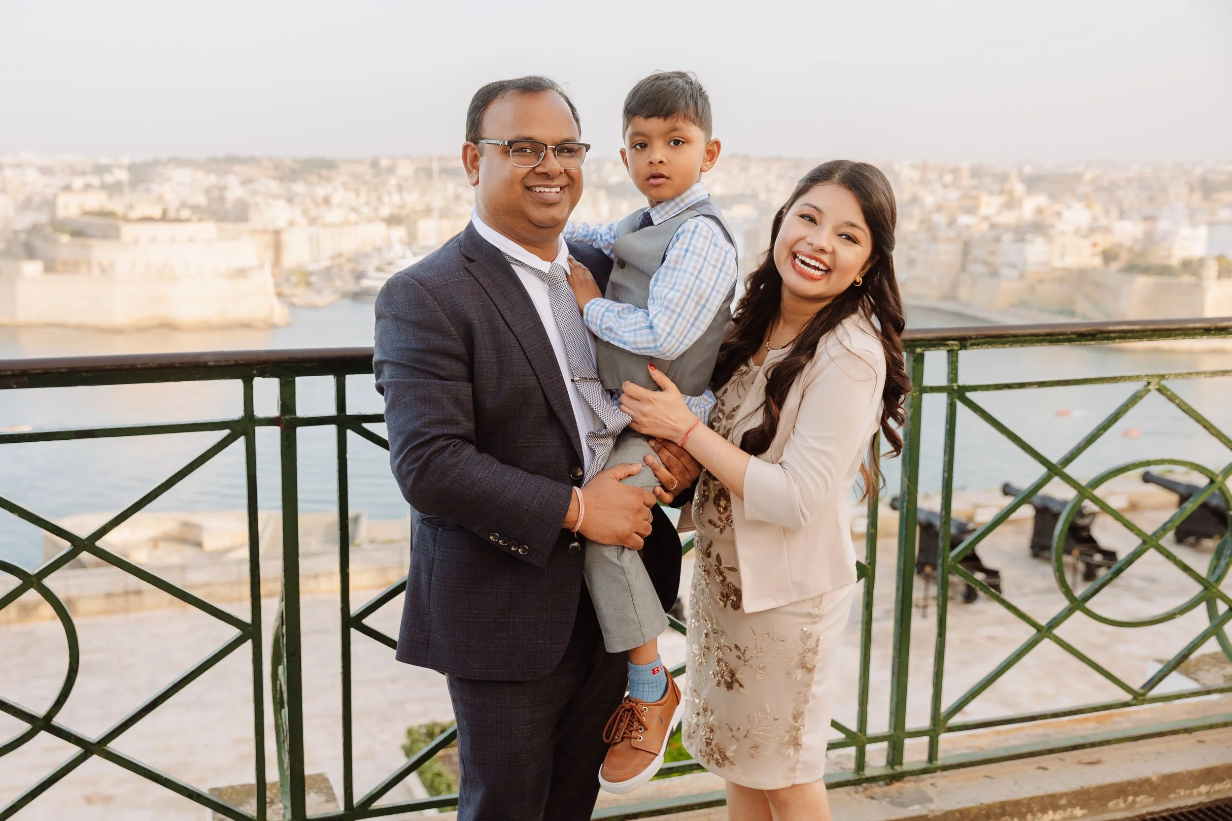 A diverse family of three smiling outdoors on a balcony overlooking a city and river, dressed in formal attire, with the father holding their young son and the mother standing beside them.
