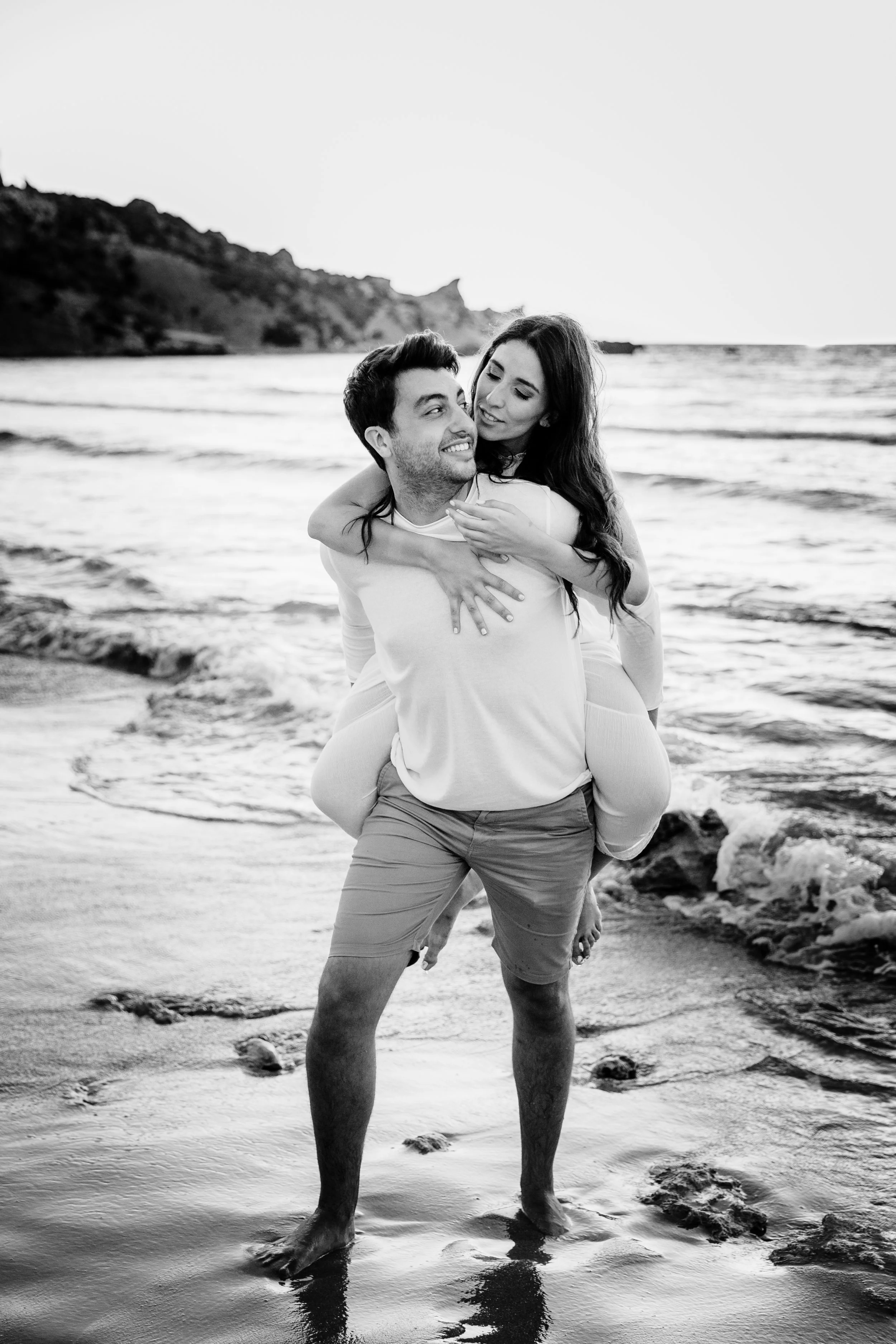 A happy couple on the beach, with the woman on the man's back, both smiling and enjoying the moment near the shoreline.