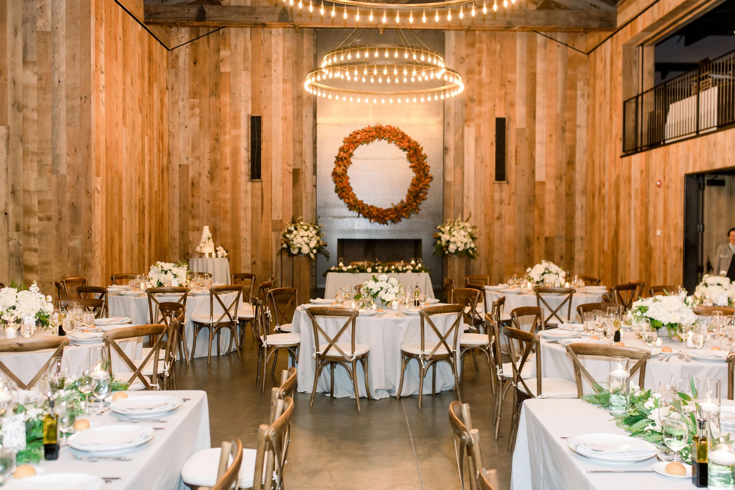 Full reception room at Brasswood Estate with round tables, white florals, wood chairs, and statement chandeliers.